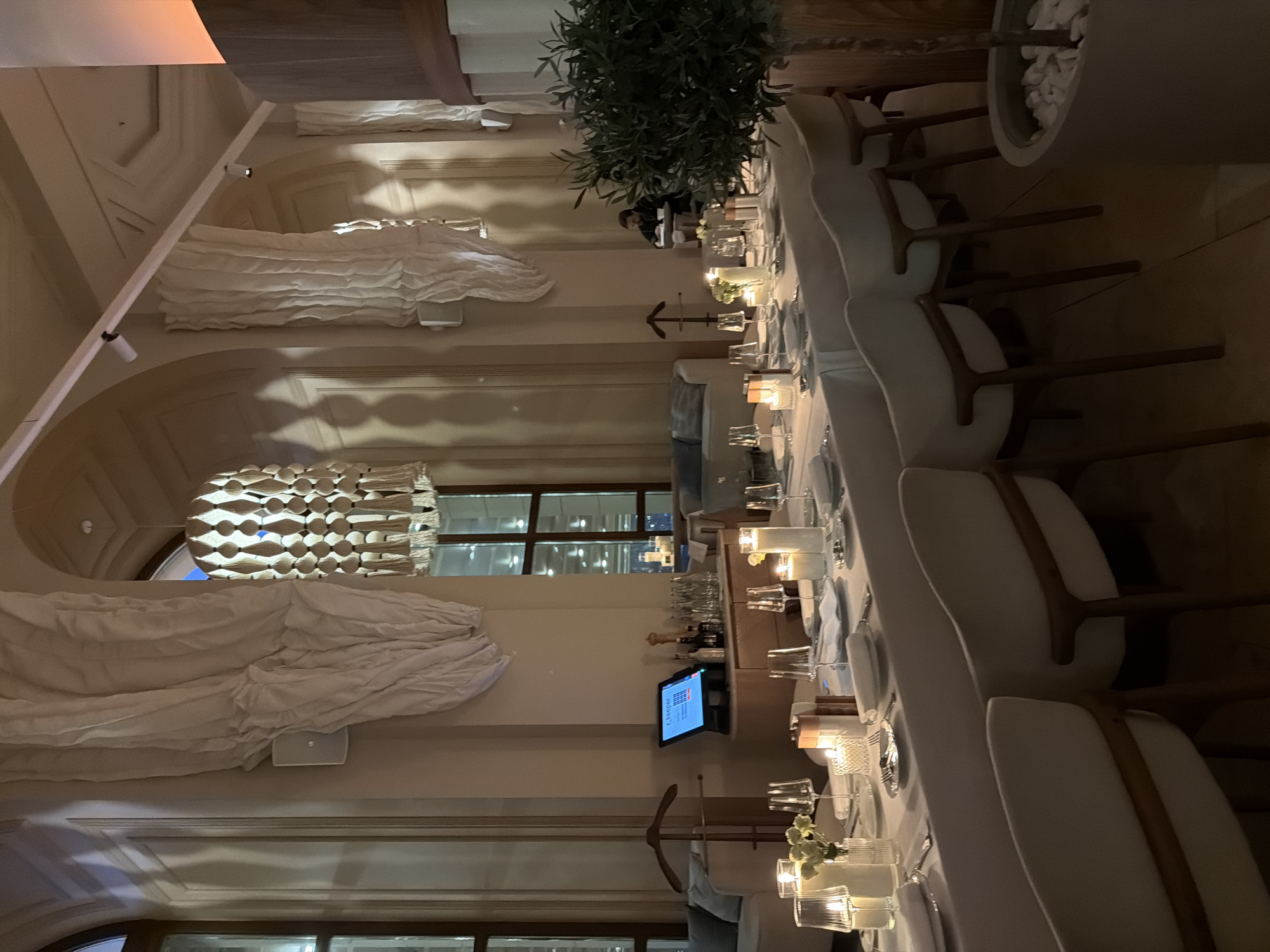 The image shows the interior of a restaurant, likely in Moscow, Russia, set up for a private event. The long table is covered with a white tablecloth and set with glassware, plates, silverware, and small candles. Several white chairs are arranged around the table. A large decorative plant is placed at the head of the table. The room has high ceilings, decorative moldings, and a large chandelier. There is a television screen behind the table displaying a menu, and some individuals are behind the plant at the head of the table.