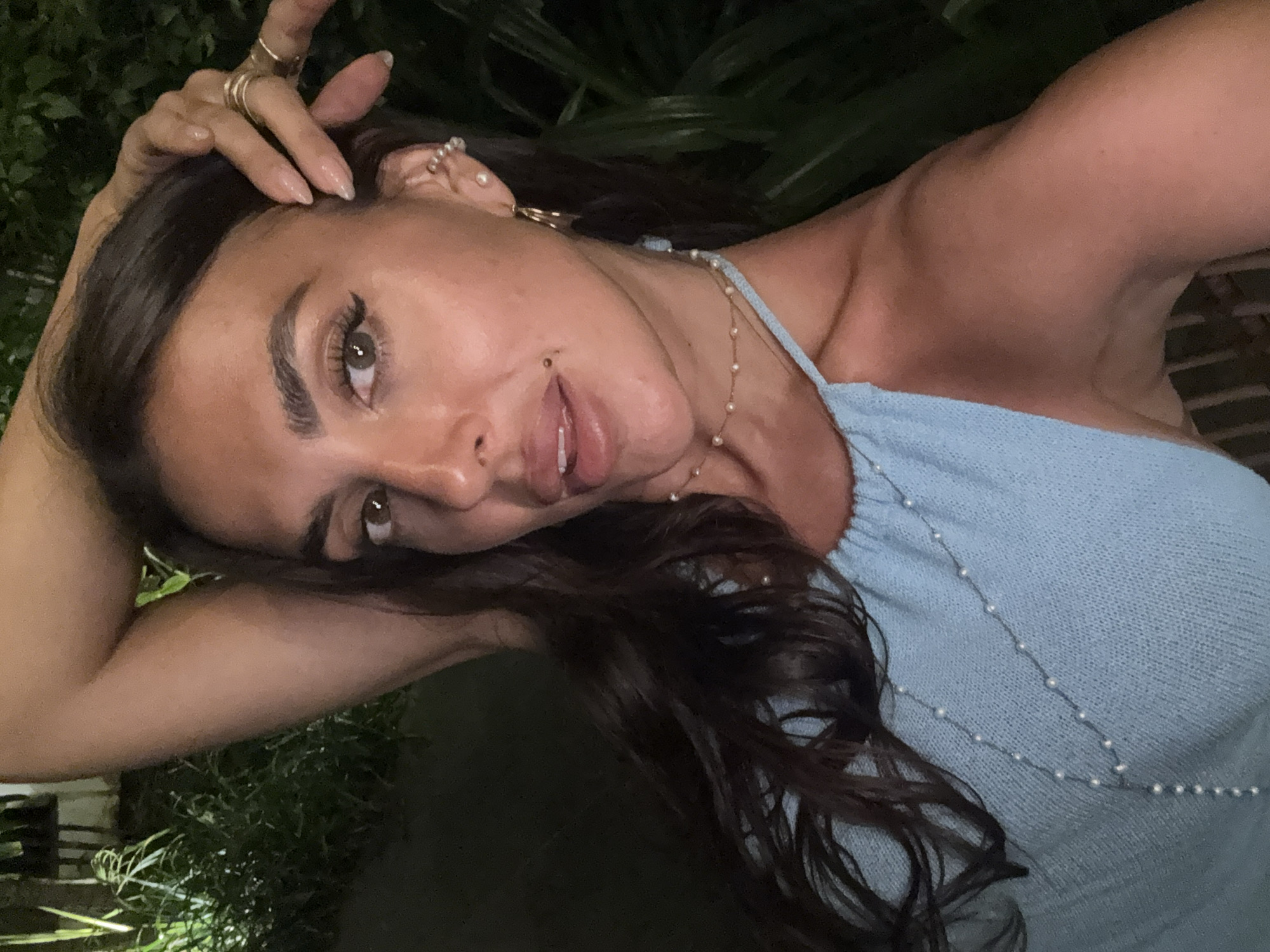 A medium shot depicts a woman with brown hair, brown eyes, and fair skin looking at the camera. She has several rings on her fingers and piercings in her ear. Her hair is styled with loose waves. She is wearing a light blue knit halter top adorned with small pearls. Her left hand rests on her head. The background features green foliage, suggesting an outdoor setting. The location is specified as Kuta Selatan, Indonesia.