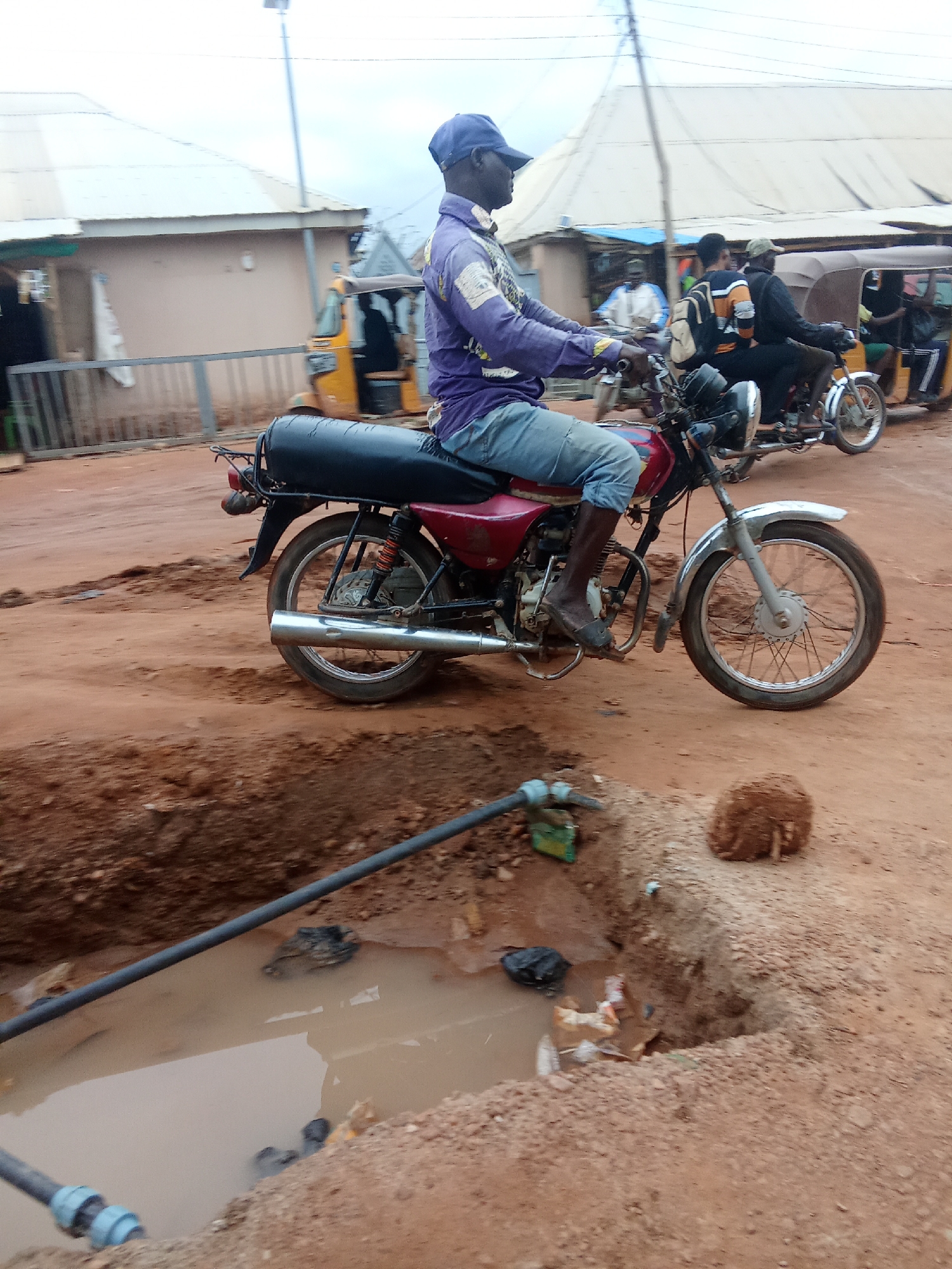 In Kagini, Nigeria, a man in a purple shirt and blue jeans rides a red motorcycle on a dirt road. He is wearing a blue cap and dark boots. Behind him, a yellow auto-rickshaw with passengers is visible, along with other motorcycles and people in the background. The road is muddy and uneven, with a large puddle of murky water and some debris, including a black plastic bag and scattered trash, on the right side of the foreground. Construction pipes are present near the puddle. Buildings with corrugated metal roofs line the street. The sky is overcast.