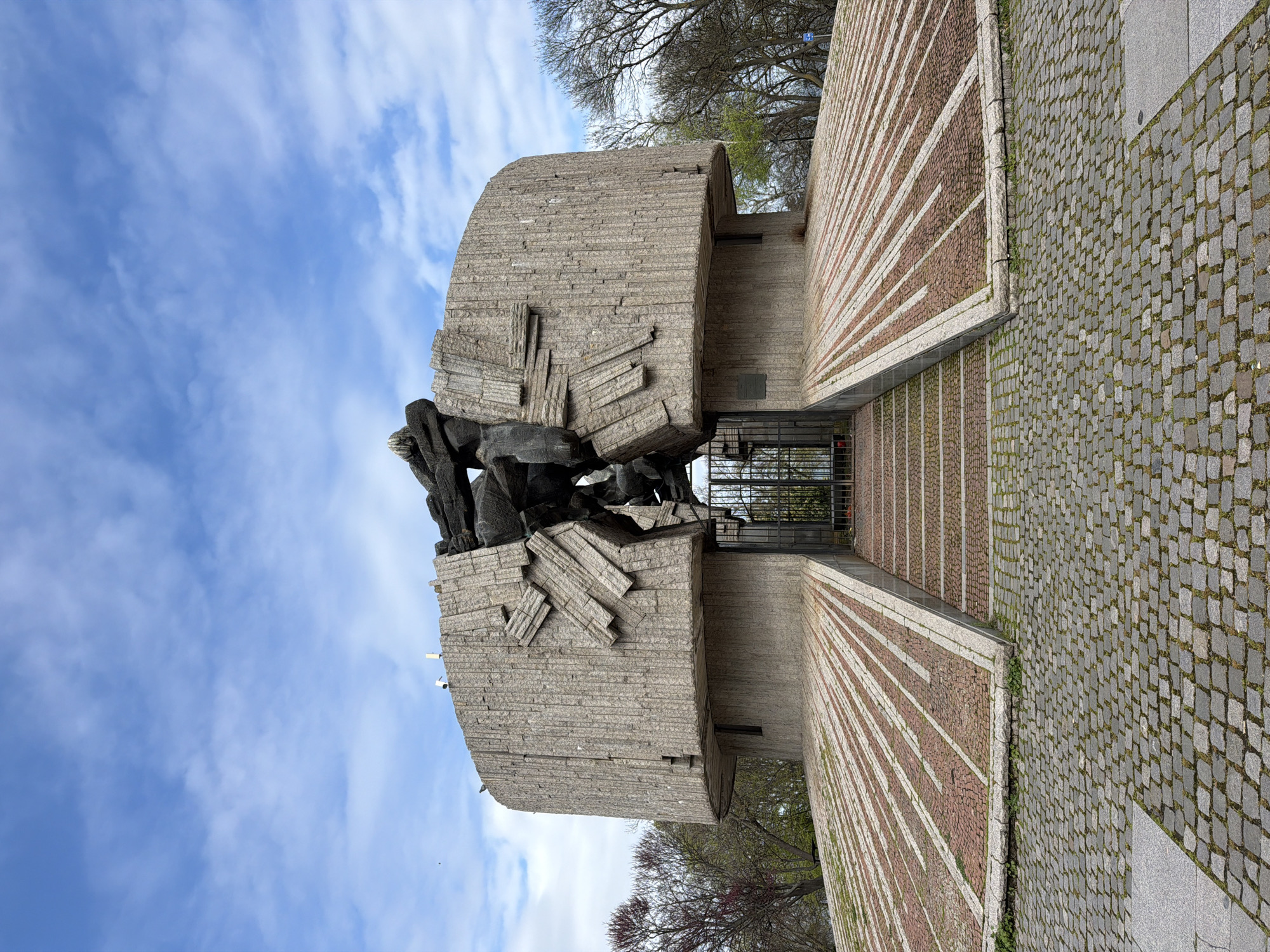 The image displays a monumental structure, likely a memorial or monument, located in Burgas, Bulgaria. The central subject is a large, imposing edifice constructed from light-grey, textured concrete or stone blocks, which gives it a layered, almost fractured appearance. Integrated into the upper section of this structure is a dark, abstract sculpture depicting several figures. These figures are rendered in a rough, blocky style, conveying a sense of struggle or dramatic action.
In front of the monument, a patterned pathway leads to an entrance marked by a dark metal gate. This pathway features prominent radiating stripes in reddish-brown and lighter grey or white, drawing the eye towards the monument's base. The surrounding ground is paved with irregular grey cobblestones, some showing small patches of green vegetation.
The scene is set outdoors under a bright blue sky with scattered white clouds, suggesting daylight conditions. Bare tree branches are visible in the background on both sides of the monument, indicating either late winter or early spring. The overall atmosphere is solemn and monumental. No specific activity is taking place, and no readable text is visible on the monument or its surroundings. The architectural style appears robust and impactful.