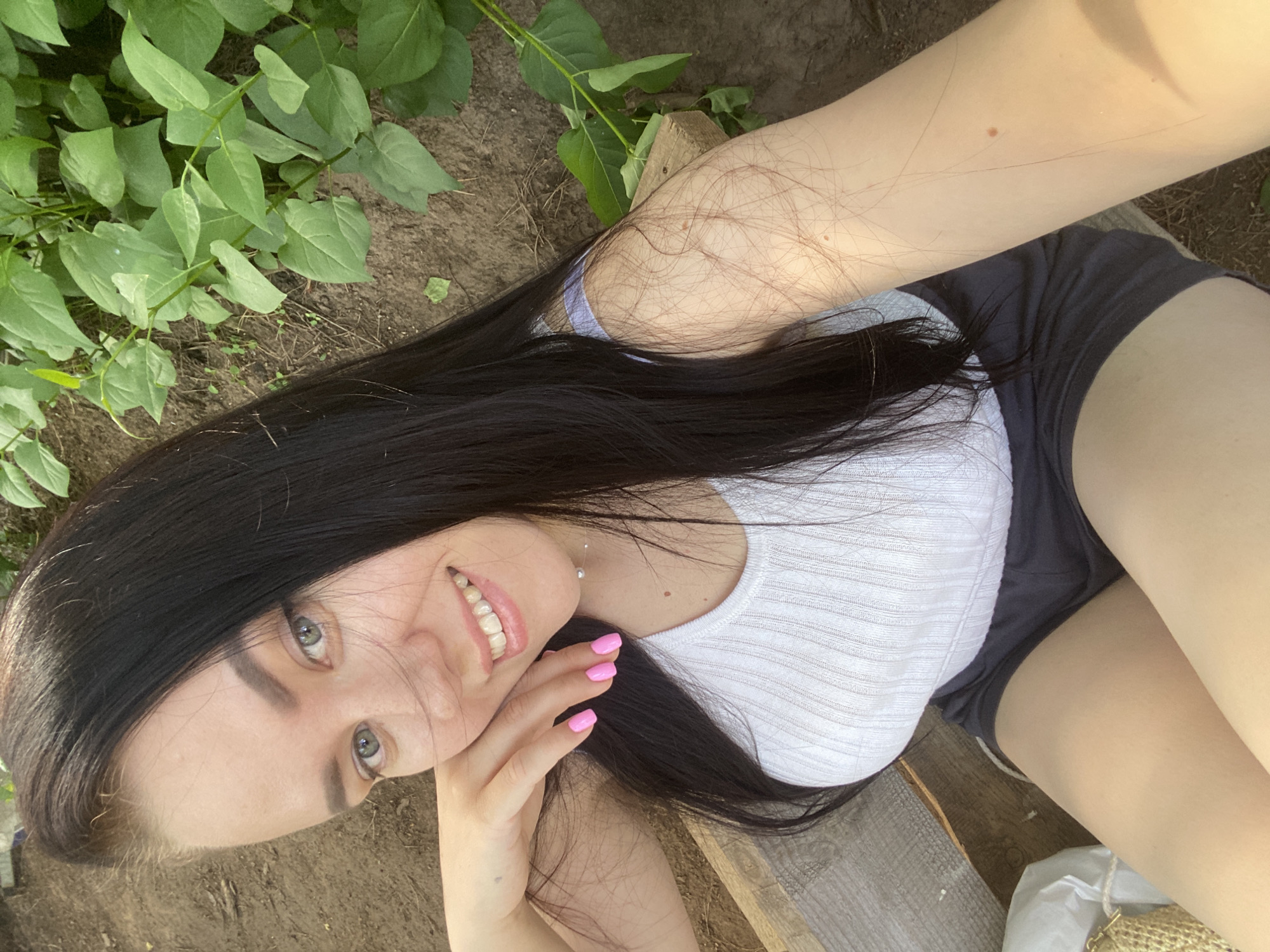 A young woman is depicted in an outdoor selfie photograph, likely taken in Chaikovsky, Russia. She has long, straight, dark brown hair, light blue-green eyes, and fair skin with visible freckles across her nose and cheeks. She is smiling, revealing her upper teeth. Her fingernails are painted a vibrant pink, and her right hand is positioned with fingers resting against her right cheek. A delicate silver necklace with a single small bead is visible around her neck. She is wearing a white, ribbed sleeveless top and dark-colored shorts with a light stripe on the left leg. She appears to be seated on a rustic wooden bench, a portion of which is visible in the lower left of the frame. To her left, a light-colored, possibly woven item is partially visible on the bench. The background consists of dark brown soil and lush green plants with broad leaves, suggesting a garden or natural cultivated area. The scene is illuminated by natural daylight.