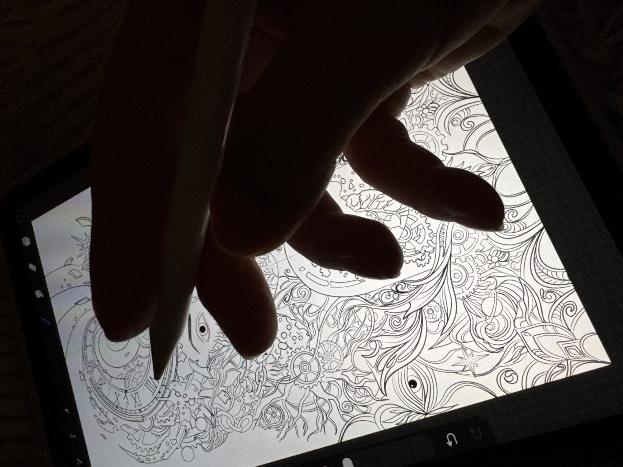 An overhead, close-up shot shows a hand holding a stylus over a tablet displaying a detailed black and white line drawing. The drawing features intricate patterns, gears, clock faces, and stylized eyes. The tablet is positioned on a dark surface, possibly a table, with the top edge of the tablet visible. The hand is silhouetted against the bright screen of the tablet, obscuring much of the drawing. The stylus is angled, poised to make a mark. The overall lighting is dim, with the tablet screen being the primary light source.