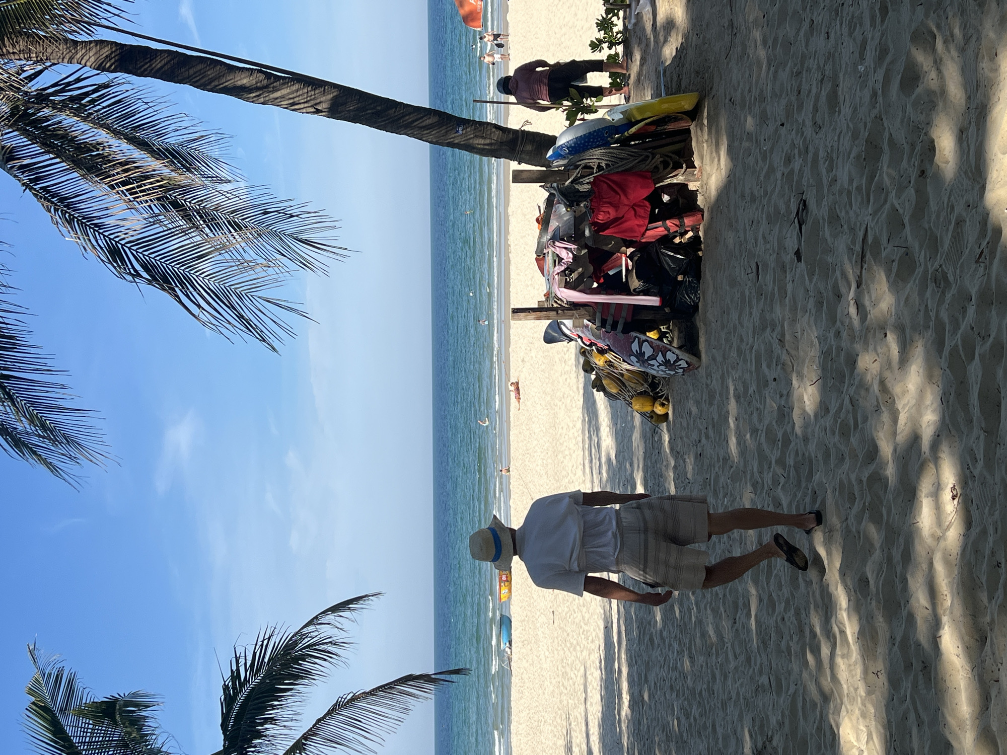 The image depicts a sunlit beach scene in Da Nang, Vietnam, likely in the late afternoon due to the long shadows cast on the sand. The foreground is dominated by sand, with palm fronds from a nearby tree partially obscuring the sky. A person wearing a light-colored short-sleeved shirt, checkered shorts, a straw hat with a blue band, and sandals stands facing away from the camera, looking towards the ocean.
Behind this person, a rack is filled with various water sports equipment, including what appear to be surfboards, kitesurfing gear, life vests in red and yellow, and coiled ropes. The ocean stretches out in the background, with gentle waves lapping the shore. Further out in the water, several figures are visible, some possibly engaged in water activities. In the distance, along the beach, a few other people can be seen enjoying the day.
The sky is a clear, bright blue, suggesting good weather. The overall atmosphere is relaxed and leisurely, typical of a beach holiday. No visible text is present in the image.