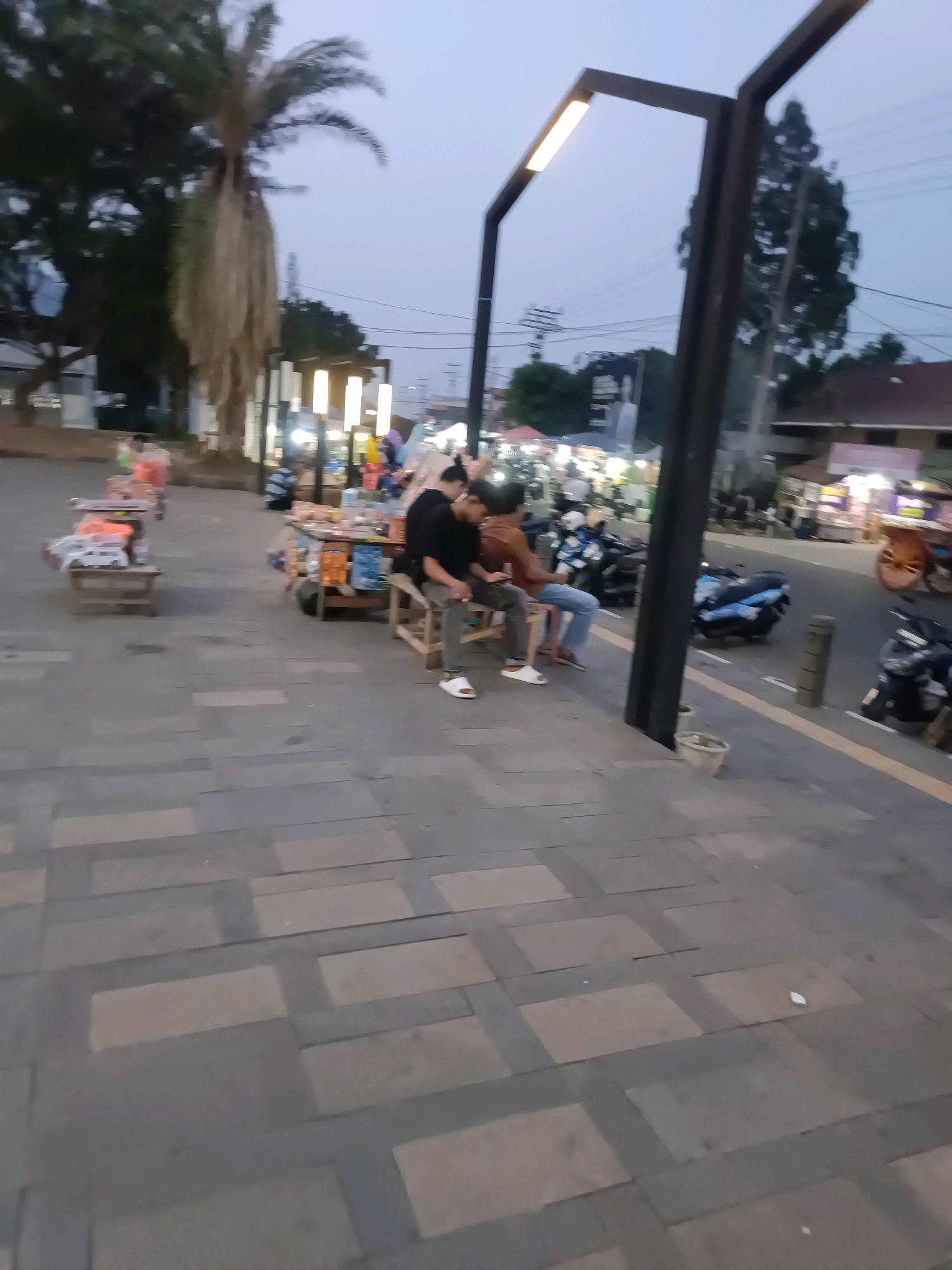 The image depicts an outdoor scene at dusk in Lubuklinggau, Indonesia, likely a public area with stalls and vendors. In the foreground, a paved walkway with a patterned surface is visible. A modern-style street lamp with a single illuminated bar stands prominently. Further back, three individuals are seated on wooden stools. Two men, one in a black t-shirt and the other in a brown shirt, are seated side-by-side. A third person, also in a dark shirt, is seated slightly apart. Behind them, several market stalls are set up, some illuminated by artificial lights, with various items displayed. Motorcycles are parked on the street in the background, and trees and buildings can be seen. The overall atmosphere suggests a bustling marketplace or an area for social gathering during the evening.