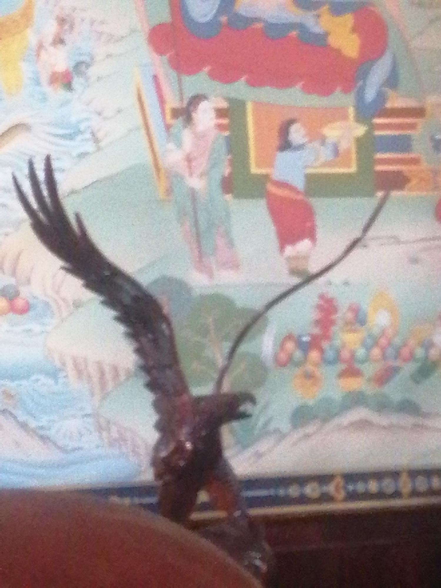 The image shows a statue of an eagle in the foreground, with a vibrant tapestry or mural in the background. The tapestry depicts stylized figures in traditional clothing, possibly from Southeast Asian or East Asian cultures, set against a landscape with mountains and abstract patterns. The eagle statue is dark brown and appears to be made of wood or a similar material, with outstretched wings. The background tapestry features bold colors like red, blue, and yellow, with intricate details and decorative elements. The scene appears to be indoors, possibly in a temple, museum, or cultural center. The overall aesthetic is rich and visually complex, with a contrast between the sculptural form of the eagle and the flat, two-dimensional artwork of the tapestry. There is no visible indication of the city of Da Lat, Vietnam within the image.
