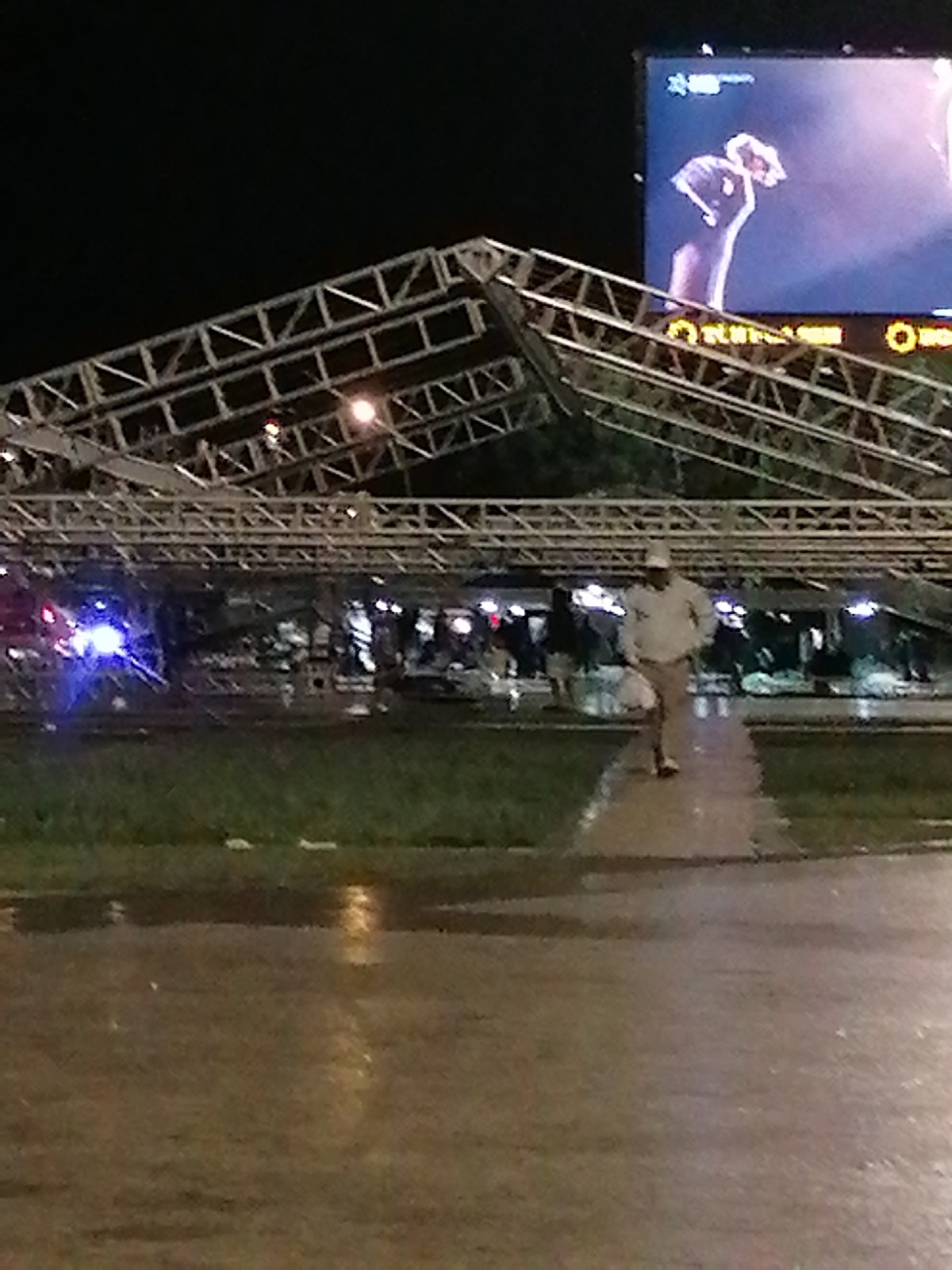 The image depicts a scene at night, likely in Da Lat, Vietnam. An elaborate metal framework structure, resembling the skeleton of a building or temporary covering, has collapsed. A man in light-colored clothing is walking away from the scene. There are multiple people in the background, some of whom appear to be standing or gathering near the collapsed structure. Police lights are visible, and a large billboard with an image on it is present in the background. The foreground shows a wet pavement, suggesting recent rain. The environment is urban.