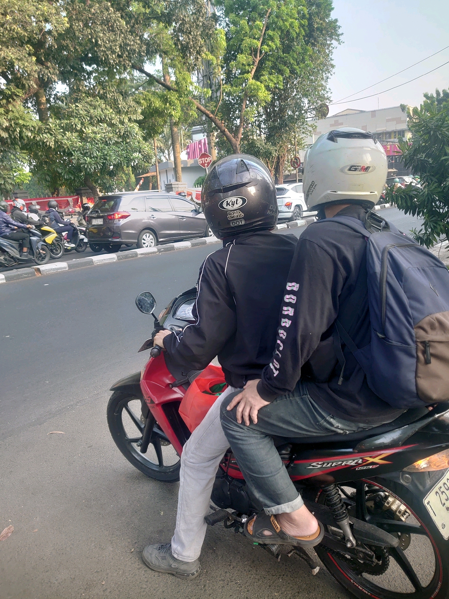 Two individuals are riding on a red and black motorcycle on a paved street in an urban environment. The person in front is wearing a black motorcycle helmet with "KYT" and "ROYAL ENFIELD" logos visible, and a black jacket. The person behind them is wearing a white and gray motorcycle helmet with a "GM" logo, and a black long-sleeved shirt with a blue backpack. The passenger has their arm around the driver. Both individuals are wearing jeans. The motorcycle has a license plate that is partially visible. In the background, there are trees, other vehicles including a gray SUV and other motorcycles, and buildings. The scene appears to be during daylight.