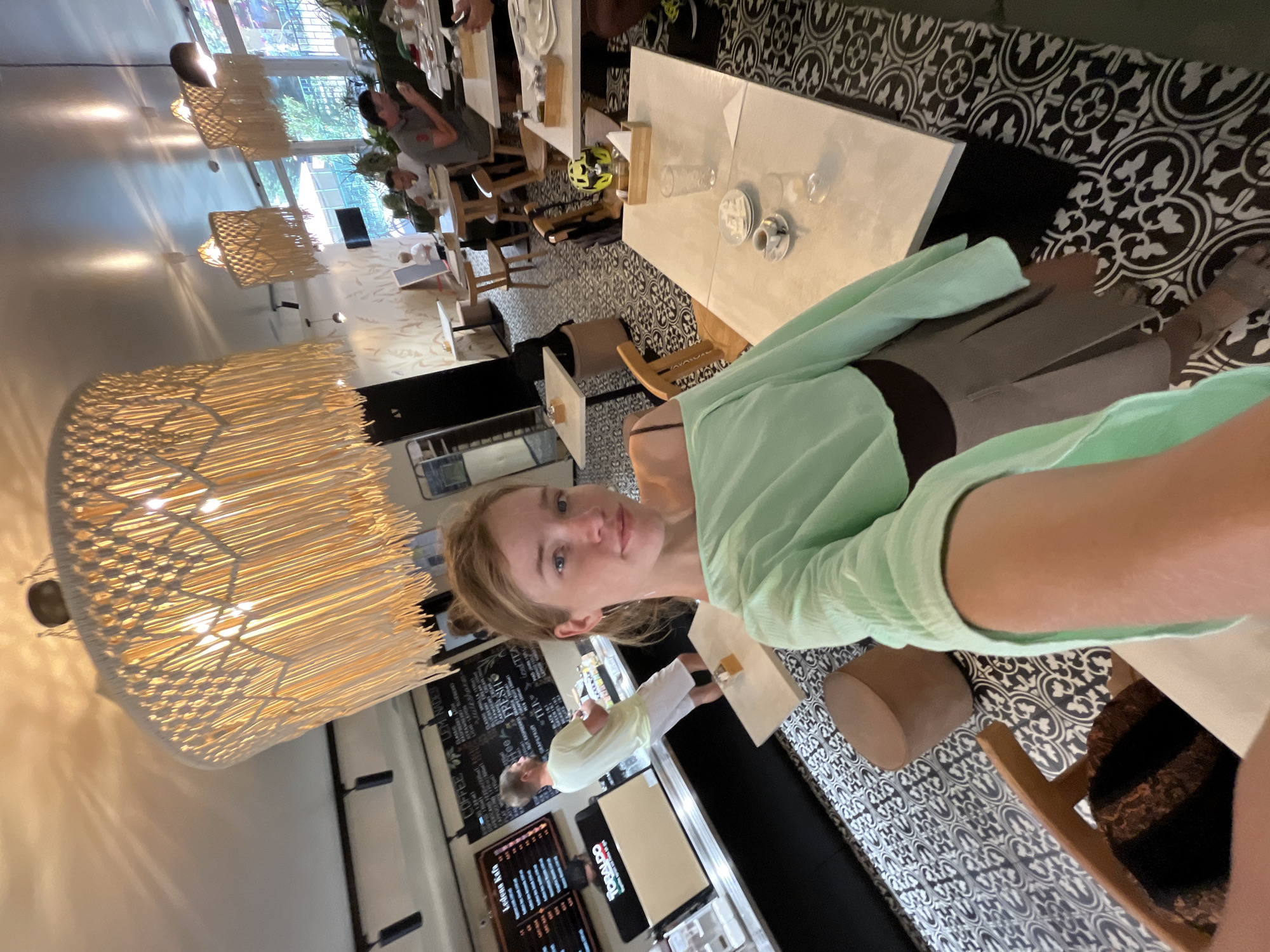 A selfie is taken by a young woman with blonde hair pulled back, wearing a light green off-the-shoulder top and a grey skirt. She is in a cafe with a distinct black and white patterned floor. In the background, a man in a light blue t-shirt and white shorts stands behind a counter with a menu board. Other patrons are seated at tables, some eating. Large, macrame-style lamps hang from the ceiling, and windows in the background offer a view of greenery. The cafe appears to be located in Lapčići, Montenegro.