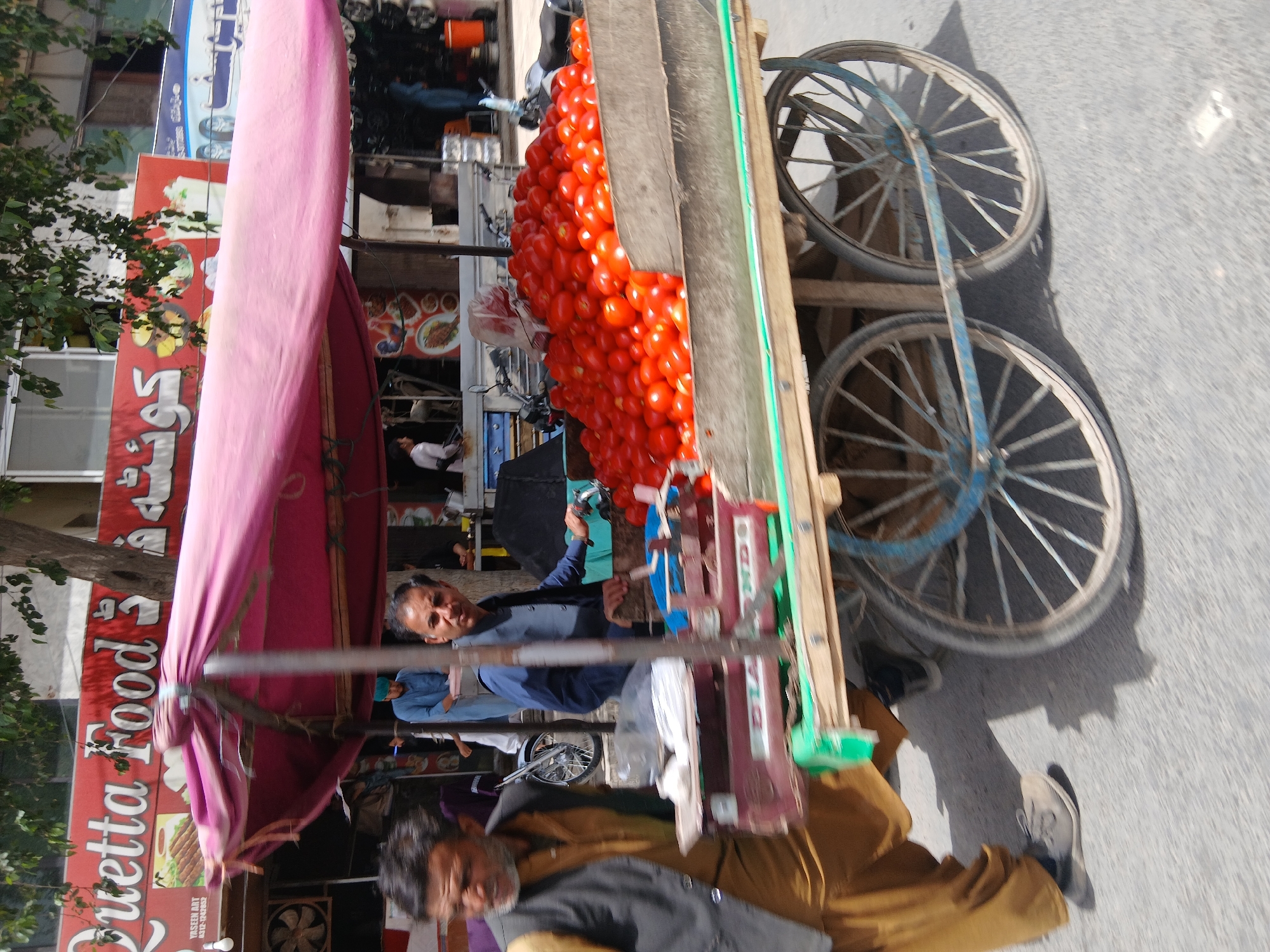 A street vendor in Quetta City Tehsil, Pakistan, is selling a large quantity of tomatoes piled on a cart. The cart has two wheels and is shaded by a pink awning. The vendor is visible, as is another person wearing a black vest and tan clothing, possibly helping with the sales. The background shows a building with signs, including one that reads "Quetta Food".