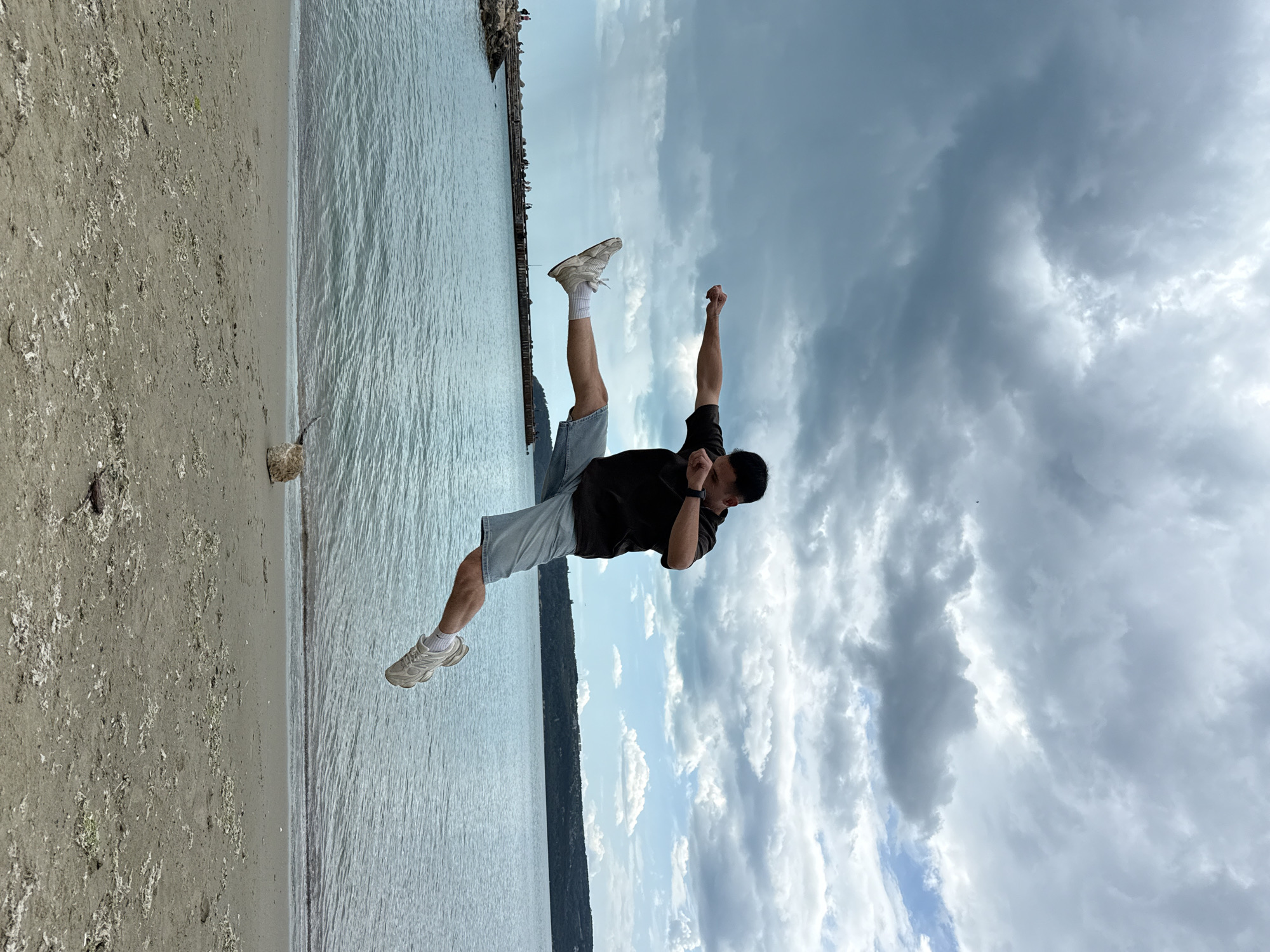 A man is in the air in a horizontal position at a beach. The beach is in Varna, Bulgaria. The man is wearing a black t-shirt, light-blue shorts, white socks, and white tennis shoes. The man is in mid-air with his legs extended and one arm bent. The other arm is extended in the air. There is a beach with water, a pier, and sand. There are clouds in the sky.