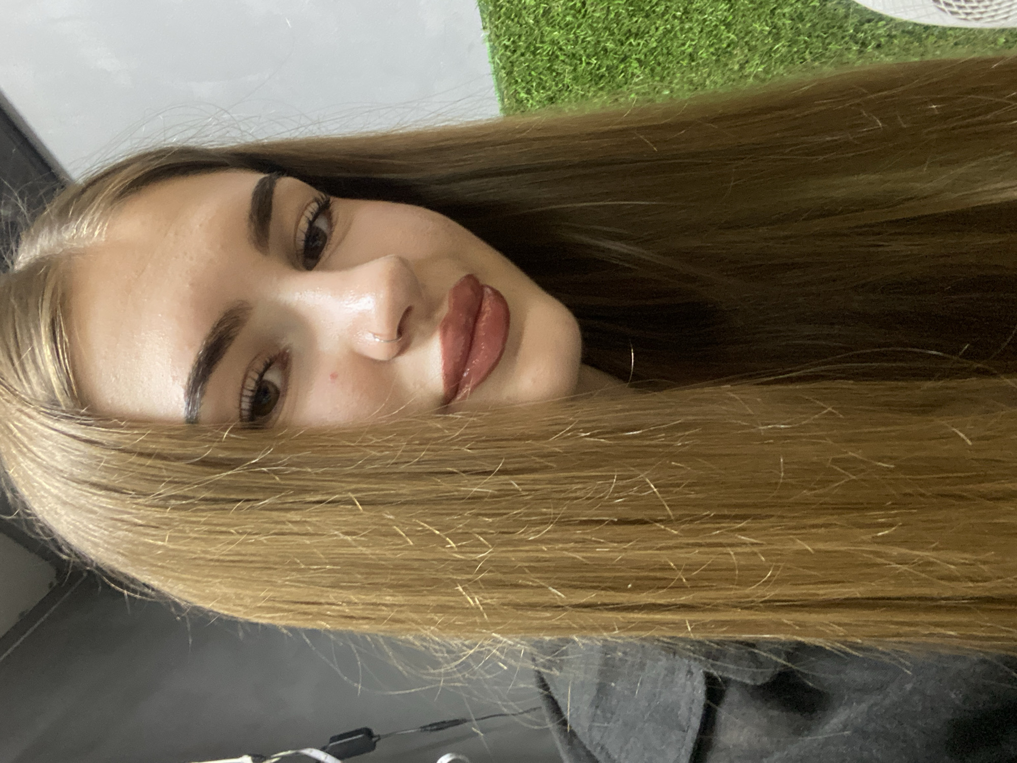 A young woman with long, straight, light brown hair is positioned in the foreground, facing slightly to her right. She is wearing a dark-colored jacket. Her face is partially obscured by her hair, but her eyes, nose with a small silver stud, and lips, which are painted with a reddish-brown lipstick, are visible. She has a light complexion and appears to be indoors. The background includes a plain grey wall on the left and a textured green surface on the right, resembling artificial turf. The scene is lit with artificial lighting. There is no indication of the specific location within Dyakovo, Russia.