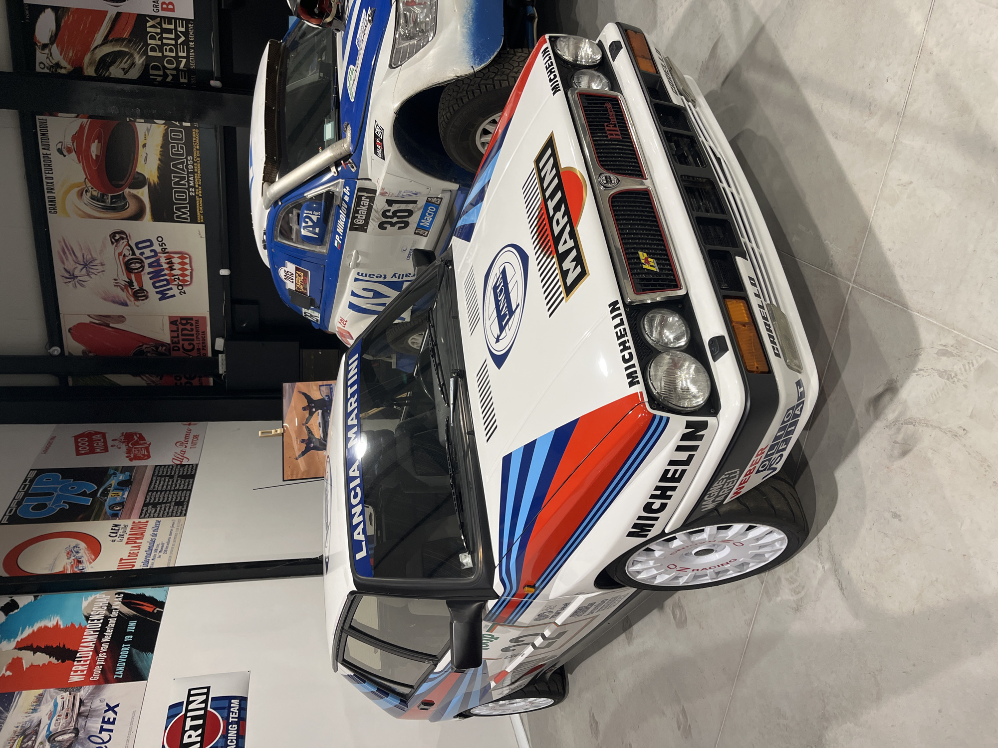 The image shows a Lancia Delta Integrale rally car with Martini livery, parked indoors on a gray tile floor. The car is white with red and blue Martini Racing stripes and Michelin tires. The front of the car is visible, including the headlights, grill, and Martini logo on the hood. Behind the Lancia is another rally car, white and blue, with the number 361 on the side. A wall behind the cars is covered with vintage motorsport posters, advertising events like the Monaco Grand Prix, the Dutch Grand Prix, and the Mille Miglia. A Martini Racing Team banner is visible at the bottom of the frame. The scene appears to be in a private collection or motorsport museum. There is no information in the image or the OCR that suggests the location is Kapatovo, Bulgaria.