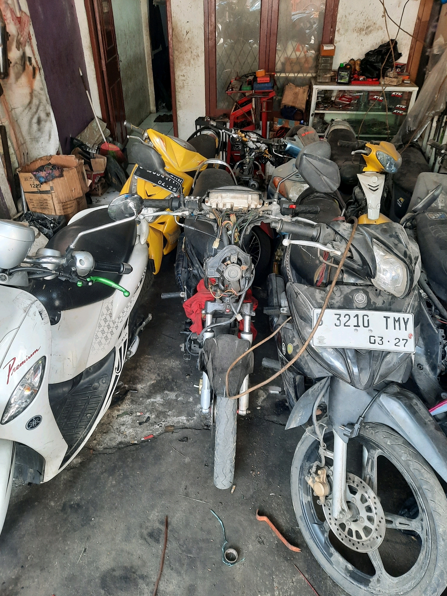 The image depicts a cluttered motorcycle repair shop in East Jakarta, Indonesia. Several motorcycles are present, including a white scooter on the left, a yellow motorcycle in the center-back, and a black motorcycle in the foreground. The black motorcycle has a license plate that reads "3210 TMY 03-27". Various tools and parts are scattered around, with some motorcycles appearing to be disassembled. Shelves filled with automotive supplies are visible in the background. The floor is concrete and appears dirty and worn, consistent with a working garage environment. No people are visible in the image.