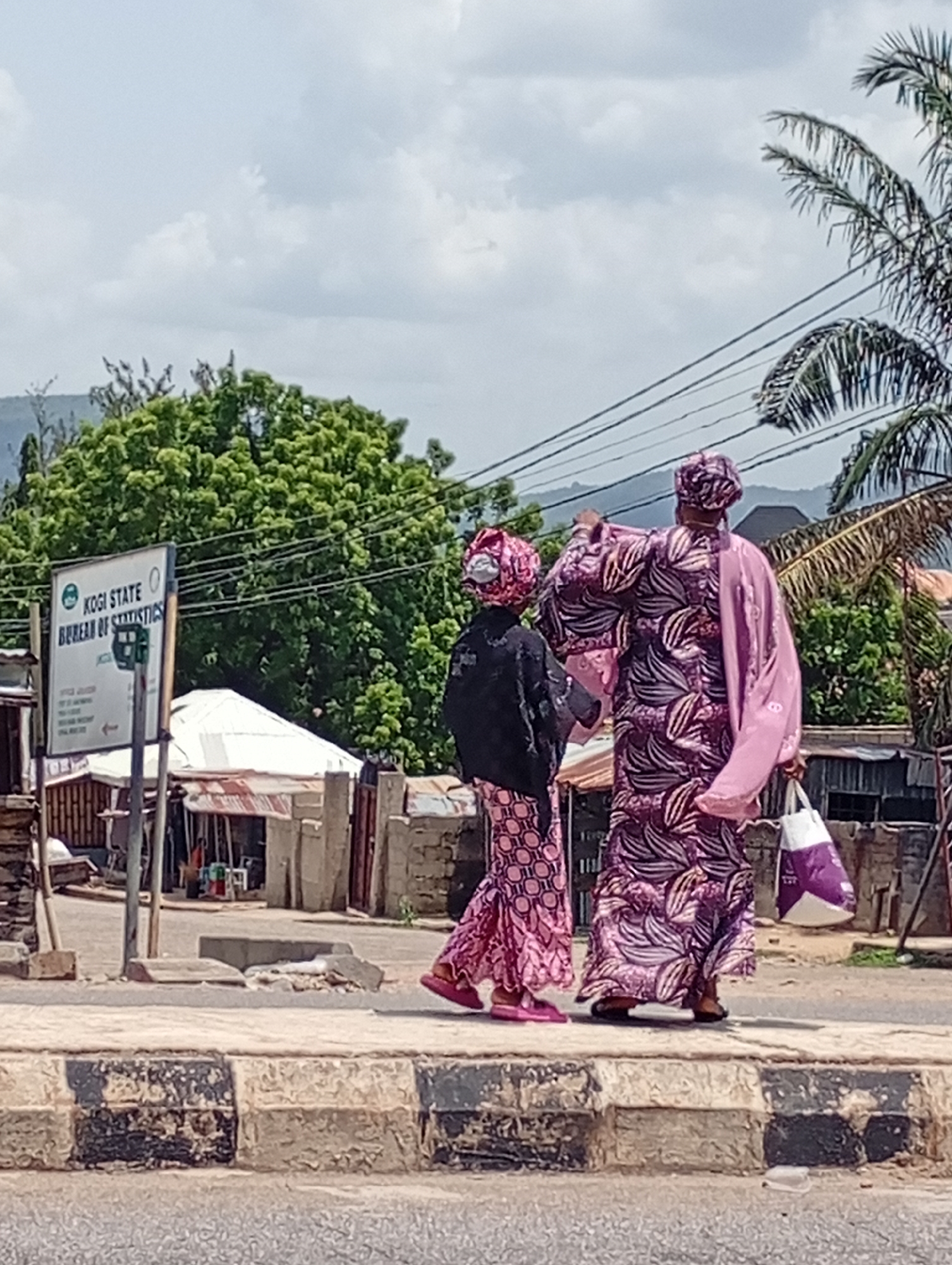 Two women are walking on a curb near a road in Unknown, Unknown. One woman is wearing a pink patterned dress with a black shawl, and a matching headwrap. The other woman is wearing a purple patterned dress with a pink shawl and a matching headwrap. She is also carrying a white and purple bag. The background includes a sign for the Kogi State Bureau of Statistics, a palm tree, a mountain, and some buildings. Power lines are visible overhead.