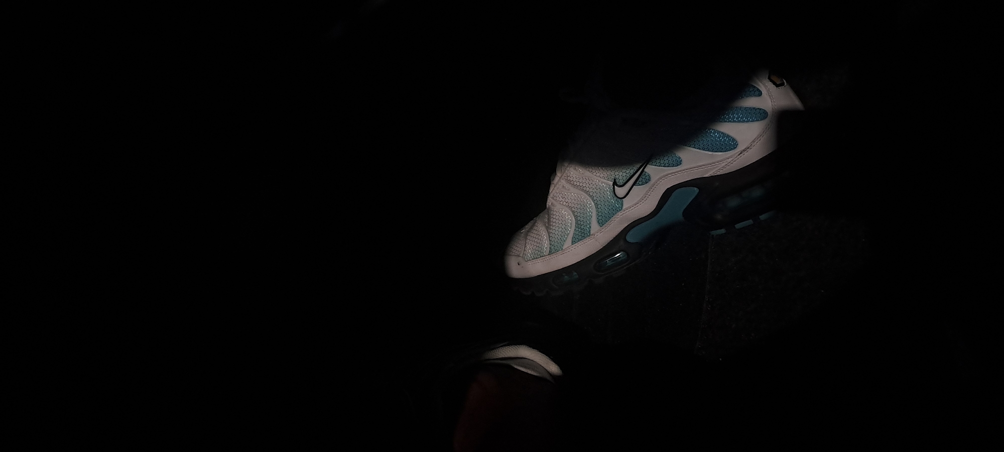 The image shows a close-up, low-angle shot of a person's foot wearing a white and light blue Nike Air Max Plus sneaker. The shoe is positioned on a dark, textured surface, likely a car floor mat or carpeting. The shoe features a visible black Nike swoosh logo on the side and a visible air cushioning unit on the sole. The lighting is dim, with the shoe being the primary illuminated subject. The background is predominantly black, suggesting a dark environment, possibly the interior of a vehicle or a dimly lit room. The overall scene is dark and slightly abstract, focusing on the details of the footwear. There is no discernible information in the image to indicate a specific location in Burgas, Bulgaria, or any interaction between subjects.