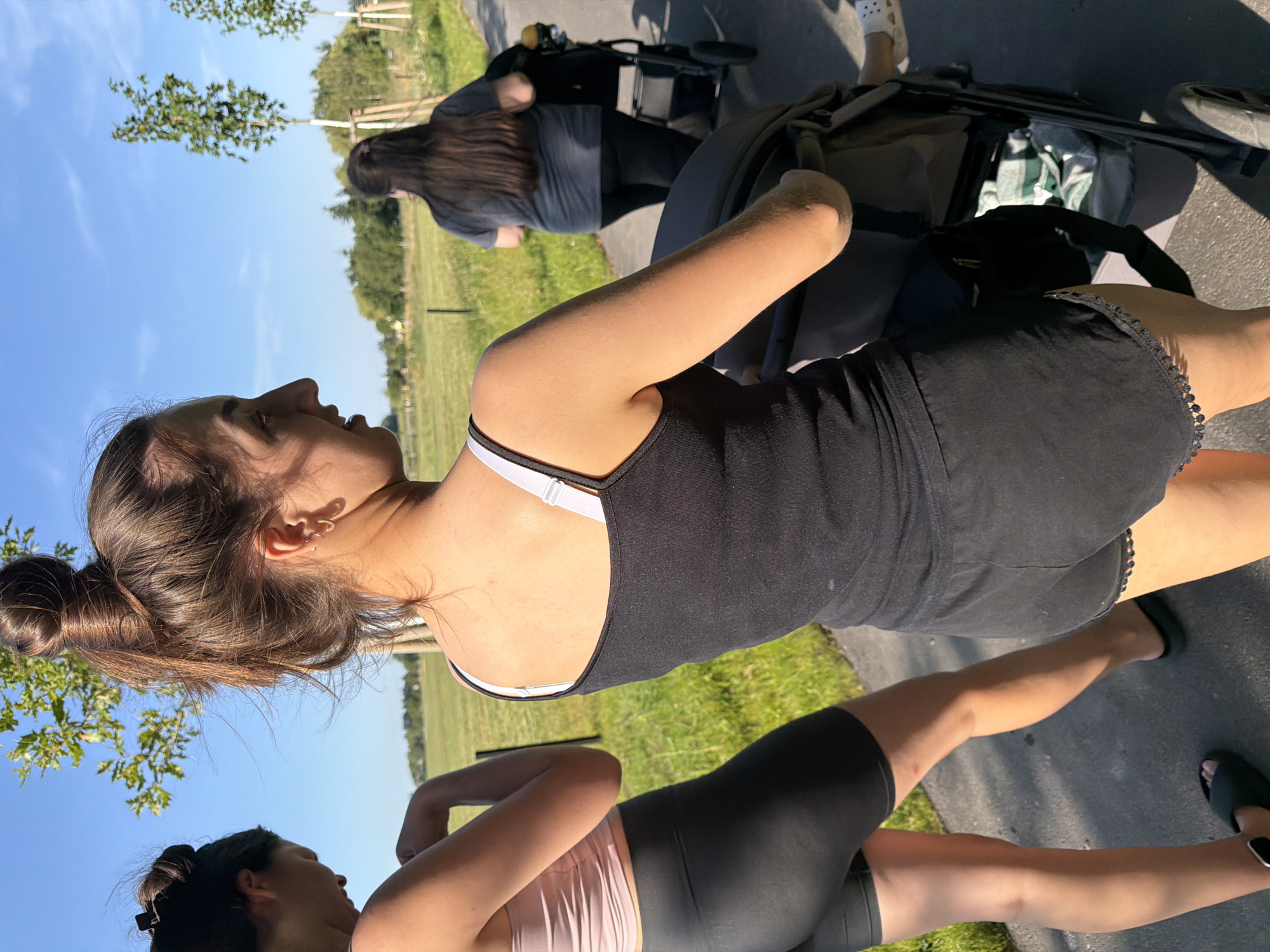 Two young women are walking on a paved path outdoors. The woman in the foreground, facing away from the camera and to the right, is wearing a black tank top and black shorts. She has her hair in a bun and is holding the handle of a stroller, which is partially visible. The second woman, to the left and also facing away from the camera, is wearing a light pink tank top and black biker shorts. She has her hair in a bun and is looking down. A third woman is walking ahead of them, pushing a stroller. She is wearing a gray t-shirt and black pants. The background consists of a grassy field, trees, and a clear blue sky, suggesting a park or rural area.