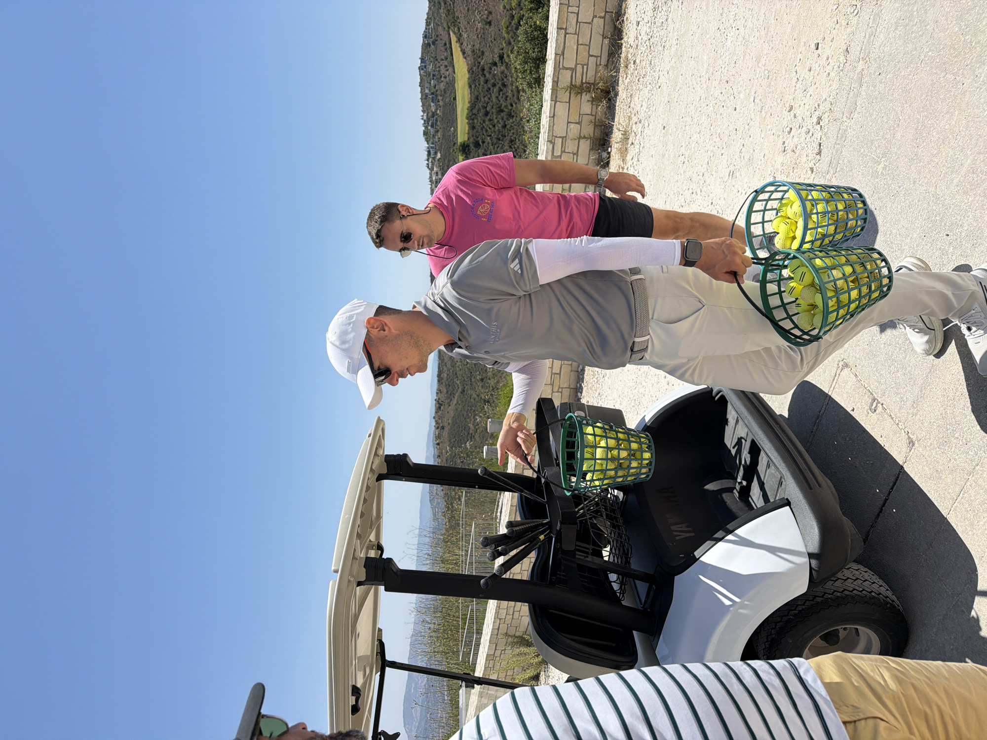 Three men are in Tsada, Cyprus, standing near a white golf cart on a sunny day. The man on the left, wearing a green and white striped shirt and tan pants, is partially visible from the side. In the center, a man in a grey polo shirt, white cap, and light-colored pants, is holding two green baskets filled with yellow golf balls. He gestures towards the golf cart with his right hand. Behind him, a man in a bright pink t-shirt and dark shorts, wearing sunglasses, stands facing away from the camera. The golf cart has several clubs in a holder. The background shows a dry, hilly landscape with some buildings visible on the slopes under a clear blue sky.