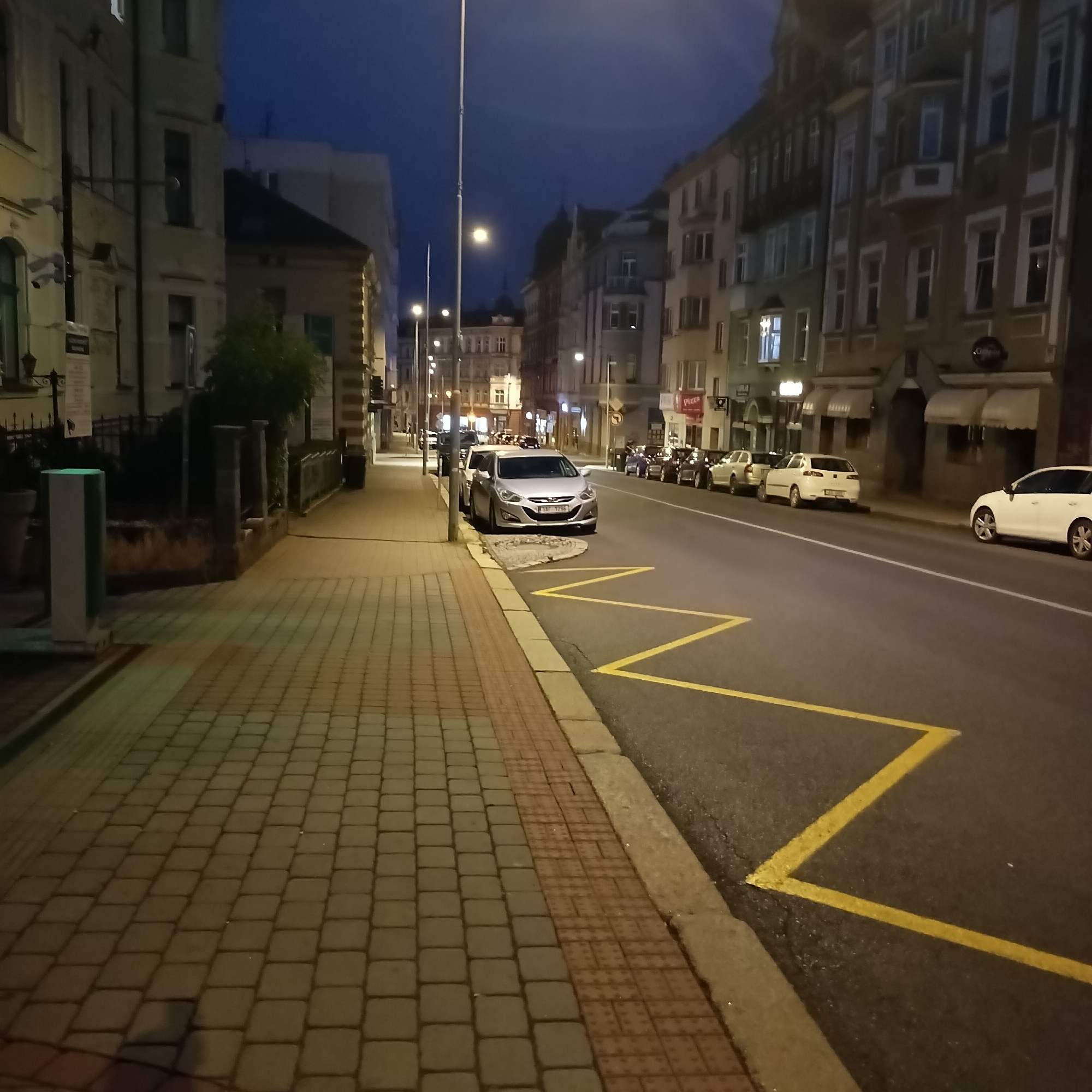 The image depicts a street scene in Cheb, Czechia, at night. The perspective is from a sidewalk, looking down a street lined with multi-story buildings. The sidewalk is paved with interlocking bricks, some of which are textured for tactile paving. A light-colored pillar is visible on the sidewalk in the foreground to the left. Across the street, several cars are parked along the curb. Yellow zigzag lines are painted on the road, indicating a no-parking zone. The streetlights and lights from the buildings illuminate the scene, casting a warm glow. The sky is a deep blue, suggesting twilight or early night. The architecture of the buildings appears to be historic, with ornate facades and numerous windows. A few trees are visible between the buildings and on the sidewalk. The overall impression is a quiet urban environment after dark.