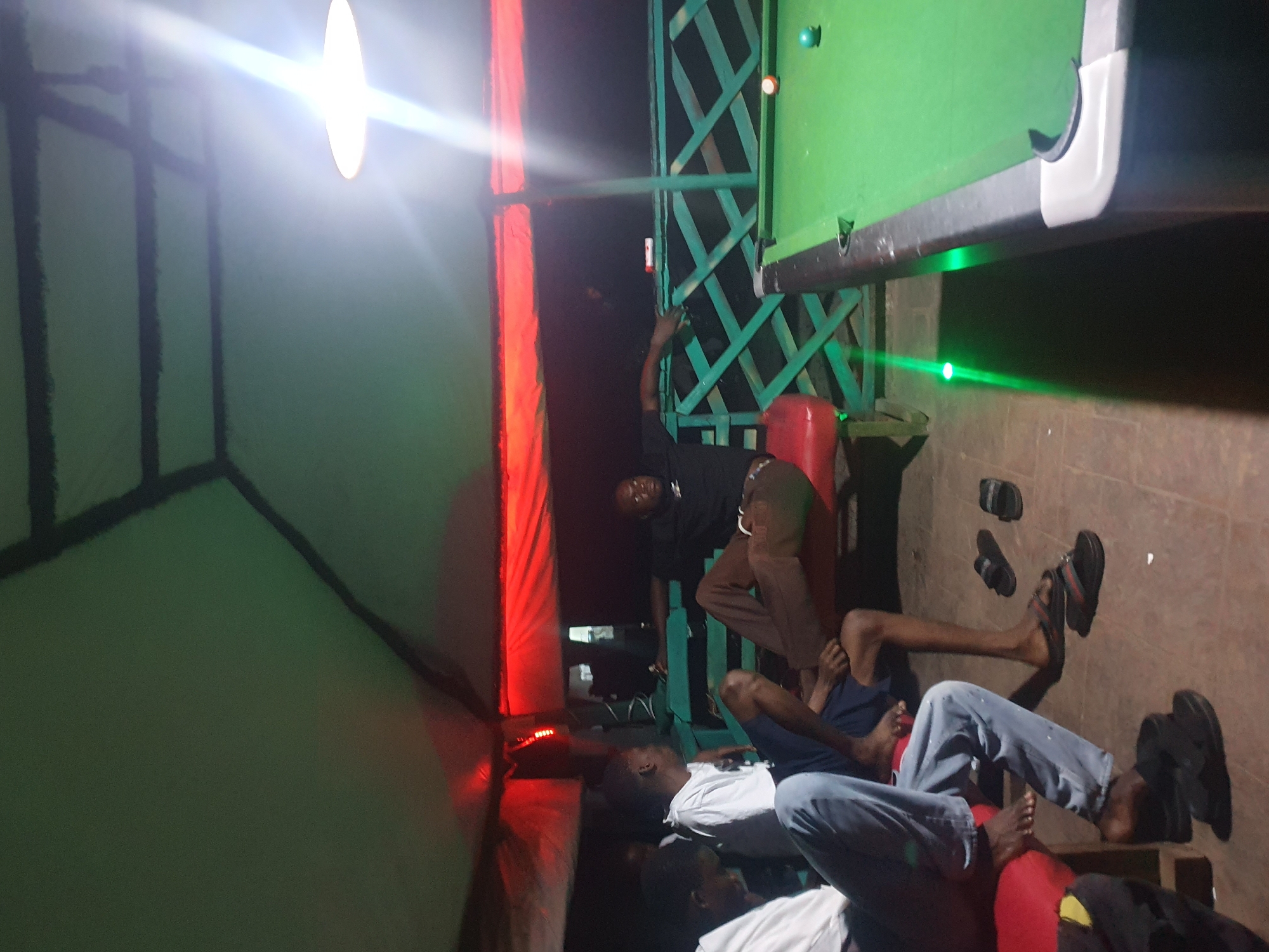 A group of men are gathered at a rooftop bar or recreational area in Lokoja, Nigeria. The setting appears to be outdoors, with a green tarp or roofing material overhead, supported by a structure with dark beams. Red lights are visible along the edge of the roofing, creating a vibrant ambiance.
In the foreground, three men are seated on a red bench. The man on the left is wearing a white t-shirt and blue jeans, facing away from the camera. Next to him, another man in a white t-shirt is looking to his right. The third man on the bench is wearing a black t-shirt and brown pants, leaning back with his arm extended. Two other men are partially visible in the background.
To the right of the bench is a pool table with a green felt surface. Several pool balls are on the table, including a cue ball and a striped ball. A green railing with a diamond pattern separates the seating area from the pool table. The floor is tiled. A bright light fixture hangs from the ceiling, casting a strong glare on the right side of the image. The overall atmosphere suggests a casual gathering or entertainment venue.