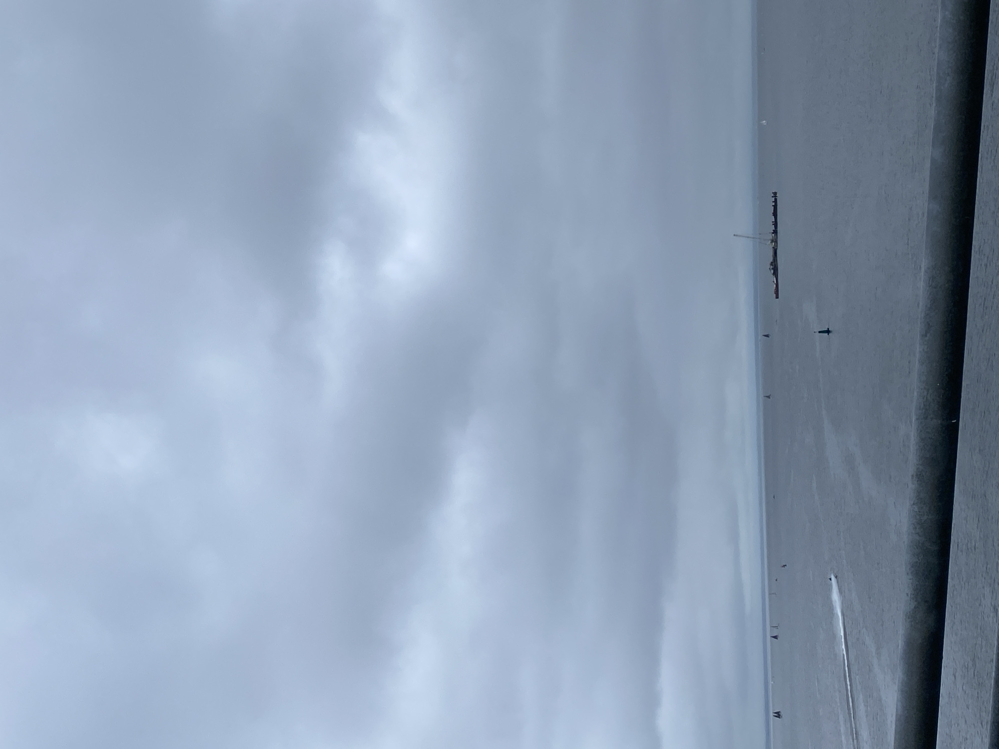 The image shows a vast, gray body of water under an overcast sky. A long pier stretches into the water, with a line of people standing at the end. Several buoys are visible in the water. There is a waterskiing boat, pulling a skier. A wooden structure occupies the right side of the frame, likely a pier or deck. The location is Saint Petersburg, Russia, as determined by the specific design of the pier.