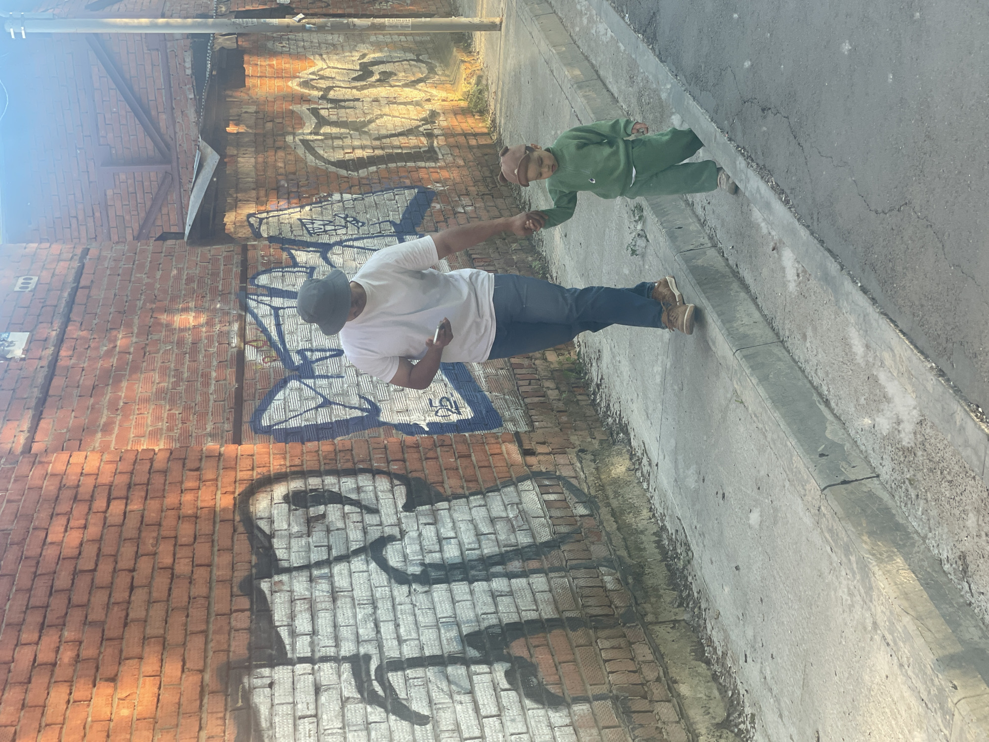 An adult male and a young child are captured in an outdoor setting on a sunny day, likely in Izhevsk, Russia. The image is taken from a slightly elevated perspective.
The adult, appearing to be a man with a dark complexion, is wearing a white short-sleeved t-shirt, dark blue jeans, and tan boots, topped with a dark grey cap. He holds a smartphone in his left hand and clasps the right hand of the child with his own right hand. The child, small in stature, is dressed in a matching green tracksuit and a light brown cap with ear-like details. The child looks towards the adult.
They are standing on a light grey paved surface, which appears to be a sidewalk or street, with a raised concrete curb running alongside it. To their left is a tall, reddish-brown brick wall, extensively covered in graffiti. The graffiti features large, stylized letters and abstract shapes rendered in white, black, and blue paint, showing signs of layering and fading in places. One particular graffiti design resembles a skull or head silhouette.
The bright sunlight suggests it is daytime, casting clear shadows and illuminating the scene. In the background, above the main brick wall, parts of another brick structure and what seem to be utility pipes are visible. The overall scene depicts a casual, everyday moment in an urban environment.