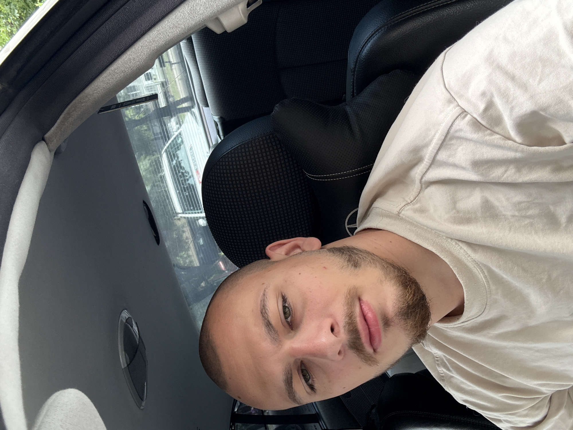 A close-up, selfie-style photo shows a young man with a shaved head and a short beard, wearing a light beige t-shirt. He is seated in the front passenger seat of a car. The background reveals the interior of the car, including a black leather headrest with stitching and a textured pattern, and the back seats. Through the car windows, trees and other vehicles are visible, suggesting an outdoor setting within a city. The lighting indicates it is daytime. Without additional context or visual cues, pinpointing the exact location within Stara Zagora, Bulgaria, is not possible.
