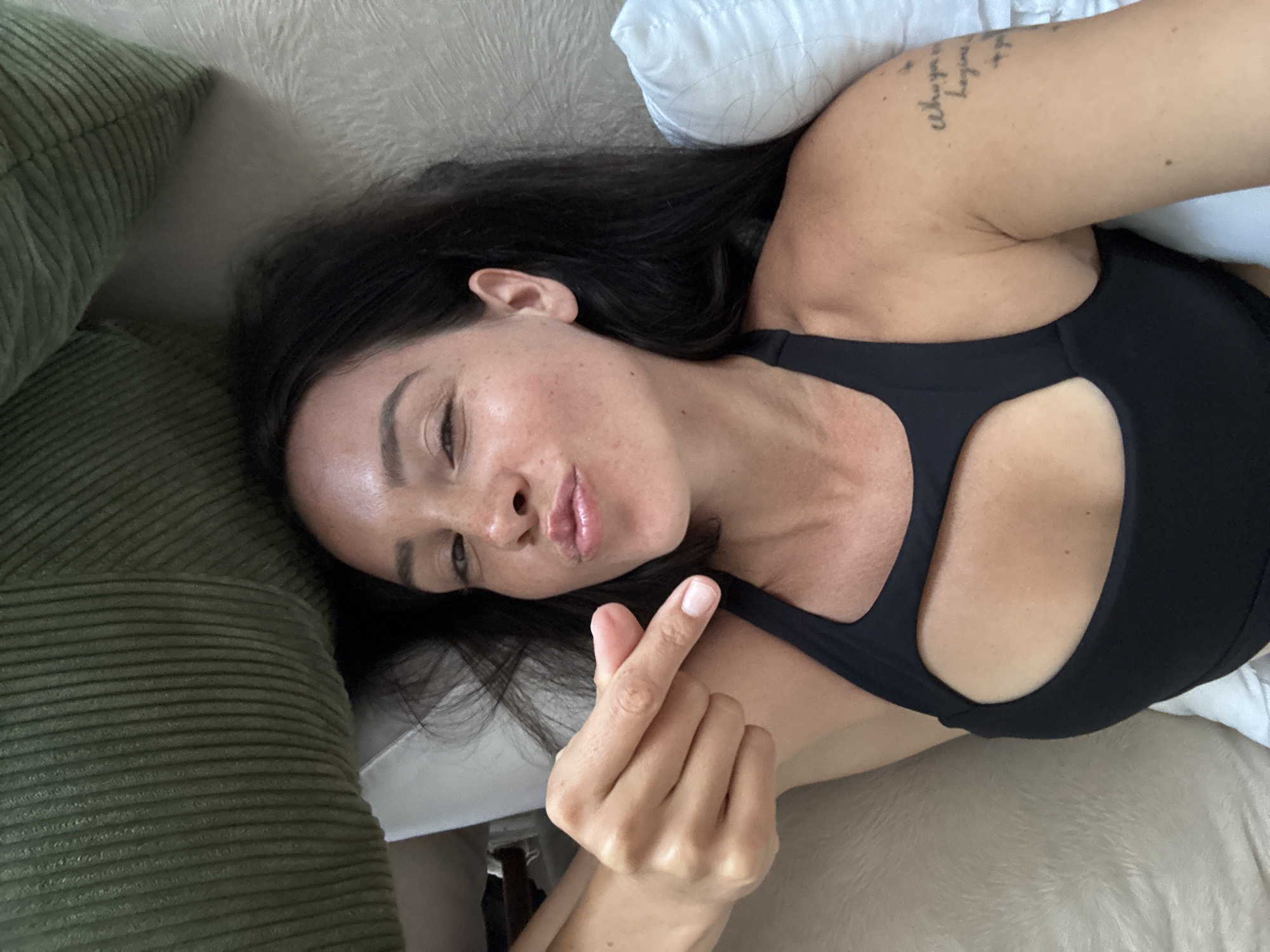 A woman with dark hair and freckles is lying down and making a kissing face. She is wearing a black sports bra with a cutout. Her left arm is bent and her index finger is extended, pointing towards her face. There is a tattoo on her right arm. She is lying on a bed with a white pillow and a green textured surface behind her. The scene is indoors, likely in a bedroom.