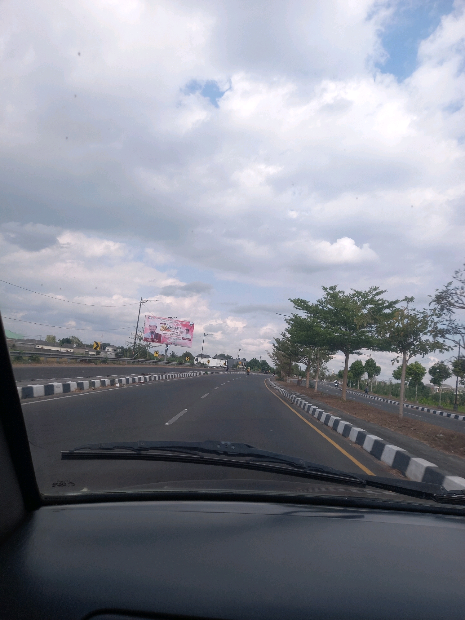 This is a photograph taken from the interior of a vehicle looking out at a road in Terong Tawah, Indonesia. The road appears to be a highway with multiple lanes and is bordered by a curb with alternating black and white markings. Along the side of the road, there are several trees, some of which are evenly spaced and appear to be ornamental. In the distance, there is a billboard with text and images on it. The sky is cloudy with patches of blue visible. The foreground of the image shows the dashboard and windshield wipers of the vehicle.