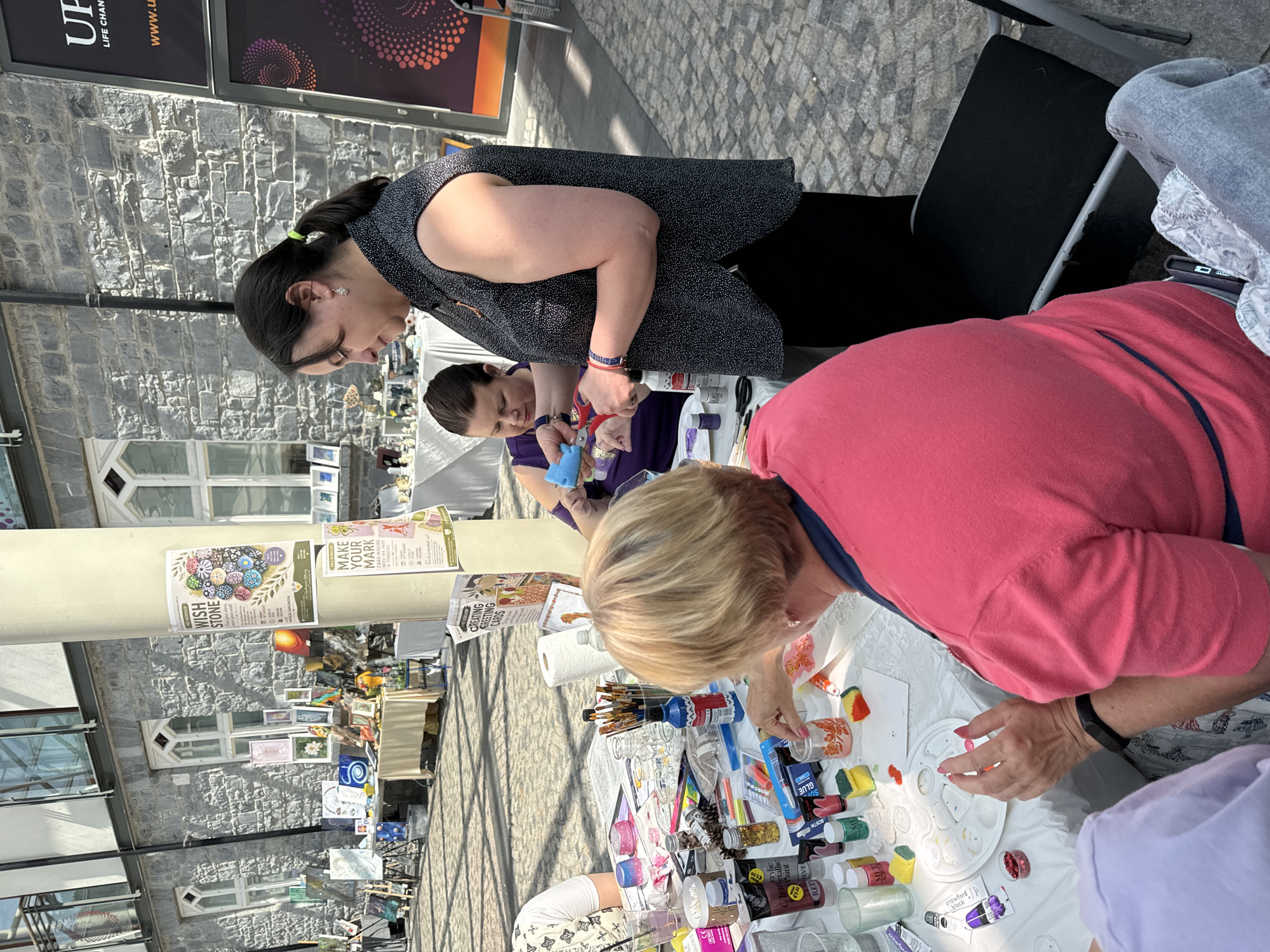 This image depicts an outdoor craft workshop taking place in Kilkenny, Ireland. In the foreground, three individuals are seated at a table laden with art supplies. A woman in a pink shirt, with blonde hair, is actively engaged in painting a small glass jar. To her left, another person, wearing a purple top, is seen holding a blue sponge and scissors, appearing to assist or participate in the activity. A third person, in a black and white polka dot sleeveless shirt, stands behind the table, holding scissors and a blue sponge, interacting with the woman in the purple top. The table is covered with a white tablecloth and is strewn with tubes of paint, brushes, small jars, glitter, glue, and other crafting materials. In the background, a stone building with arched windows serves as a backdrop, and a pillar displays posters for art workshops, including "Wish Stone" and "Make Your Mark." The ground is paved with cobblestones, suggesting a historic urban setting. Further in the background, stalls displaying artwork are visible, indicating a market or festival atmosphere. The overall scene suggests a creative and interactive community event.