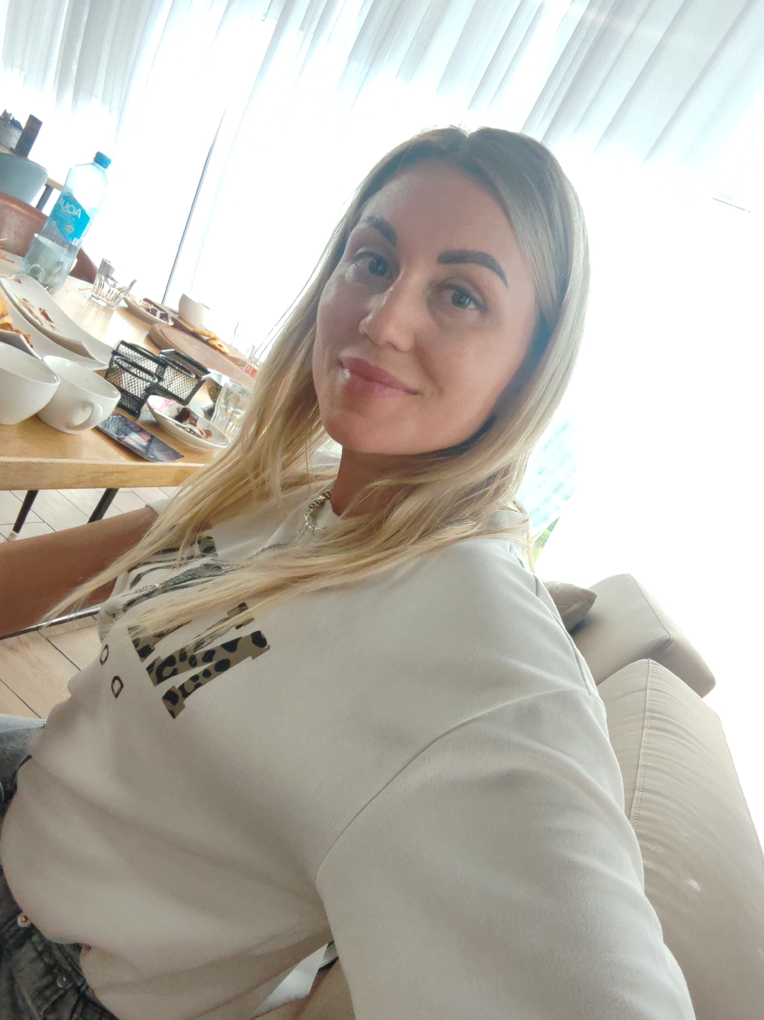 The image shows a woman taking a selfie in a brightly lit room, possibly a cafe or restaurant, with white curtains in the background and a table with food and drinks in front of her. The woman has blonde hair and is wearing a white t-shirt with a leopard print design. The table has plates with remnants of food, cups, a bottle of water, and a metal wire basket. The presence of a sofa is visible in the lower right corner of the image. The image does not provide any specific visual cues to determine the location within the city of Amursk, Russia.