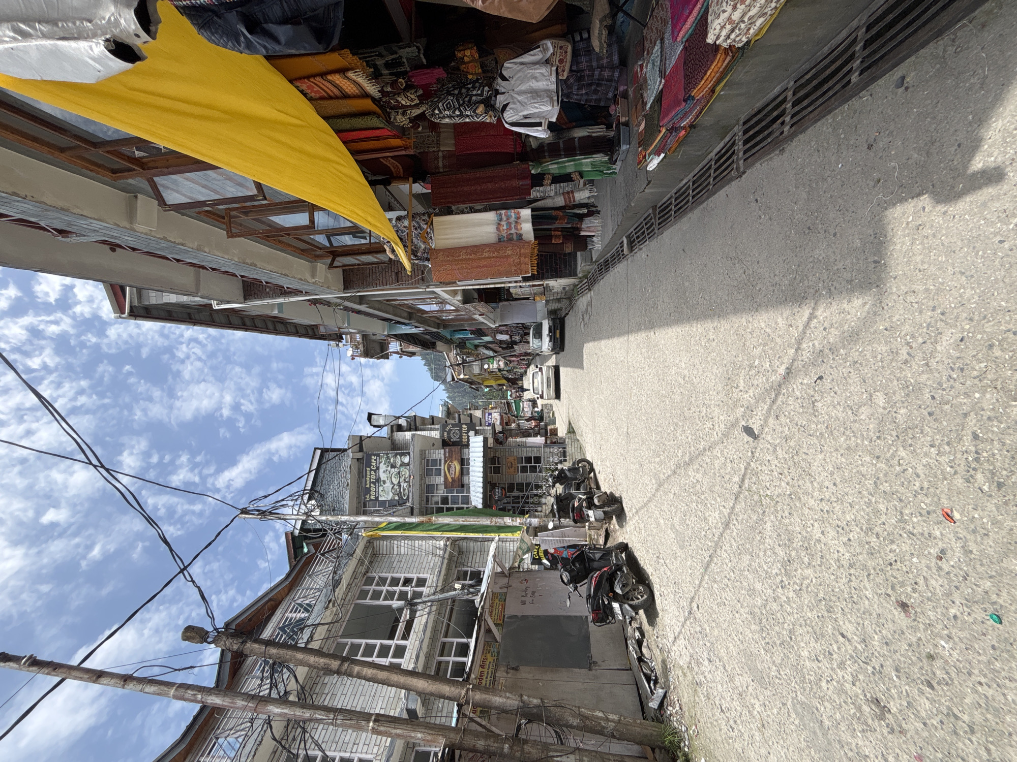 The image depicts a street scene in Vashisht, India, characterized by a narrow, paved road lined with multi-story buildings. The buildings on the left are primarily light-colored, featuring multiple windows and balconies, with visible electrical wiring and utility poles. A sign on one building reads "BACKYARD ROOF TOP CAFE". Several scooters and motorbikes are parked along the left side of the street.
On the right side of the street, shops display colorful textiles, scarves, and clothing items, with a prominent yellow tarp covering part of the storefronts. A white car is visible further down the street, as is another car approaching from the distance. The sky is blue with scattered clouds. The overall impression is of a bustling market or commercial street in a hilly town.