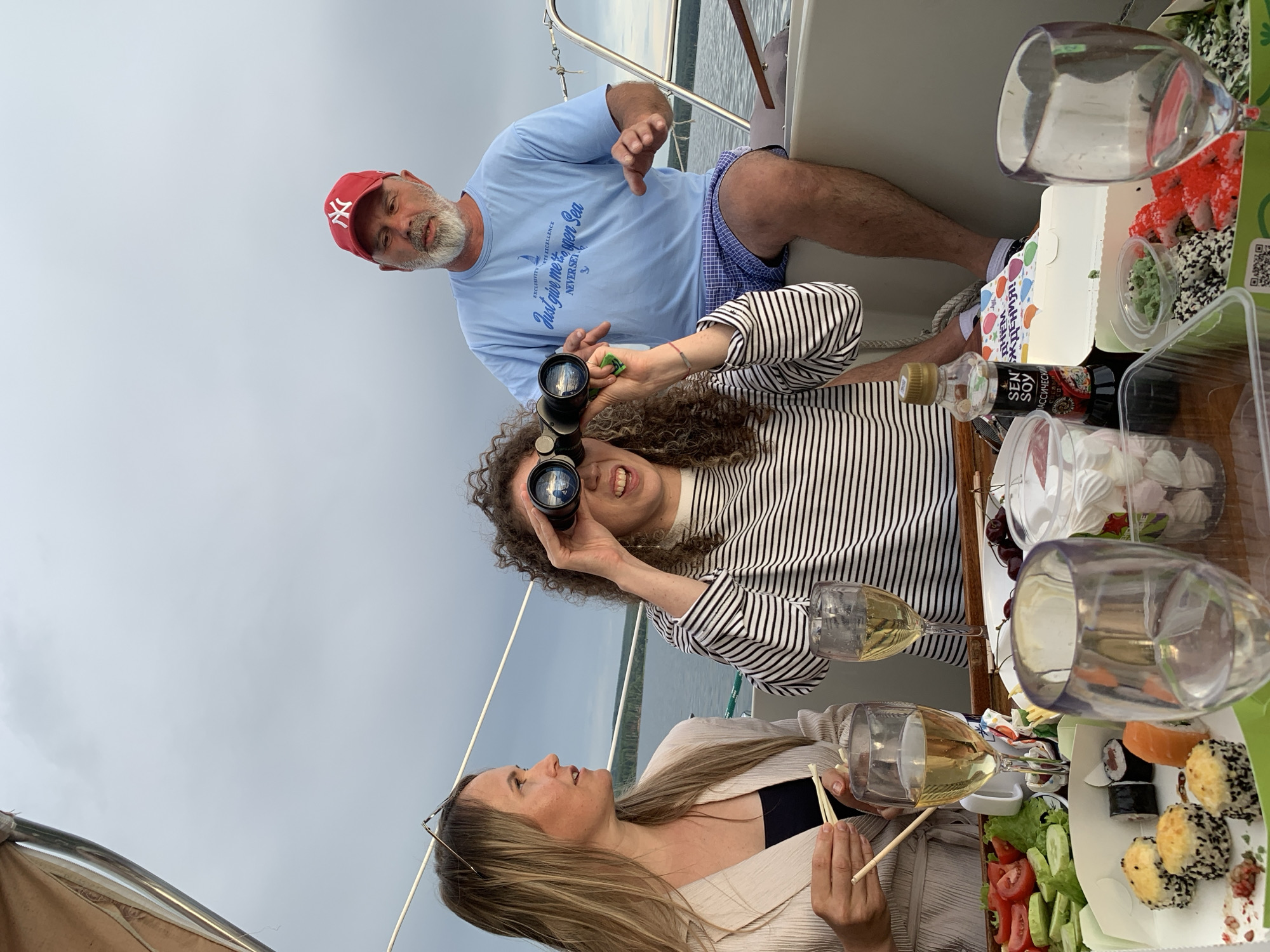 Three people are on a sailboat in Chaykovsky, Russia, with a spread of sushi and drinks on a table in the foreground. The sky is overcast. A woman with blonde hair is on the left, wearing a light-colored blazer over a black top, and holding chopsticks. In the center, a woman with curly brown hair is looking through binoculars, wearing a striped shirt. A man with a grey beard and a red New York Yankees cap is on the right, wearing a light blue t-shirt and blue plaid shorts. He is looking to his left and gesturing with his hands. The table has wine glasses, a box of sushi, a salad, and a bottle of soy sauce.