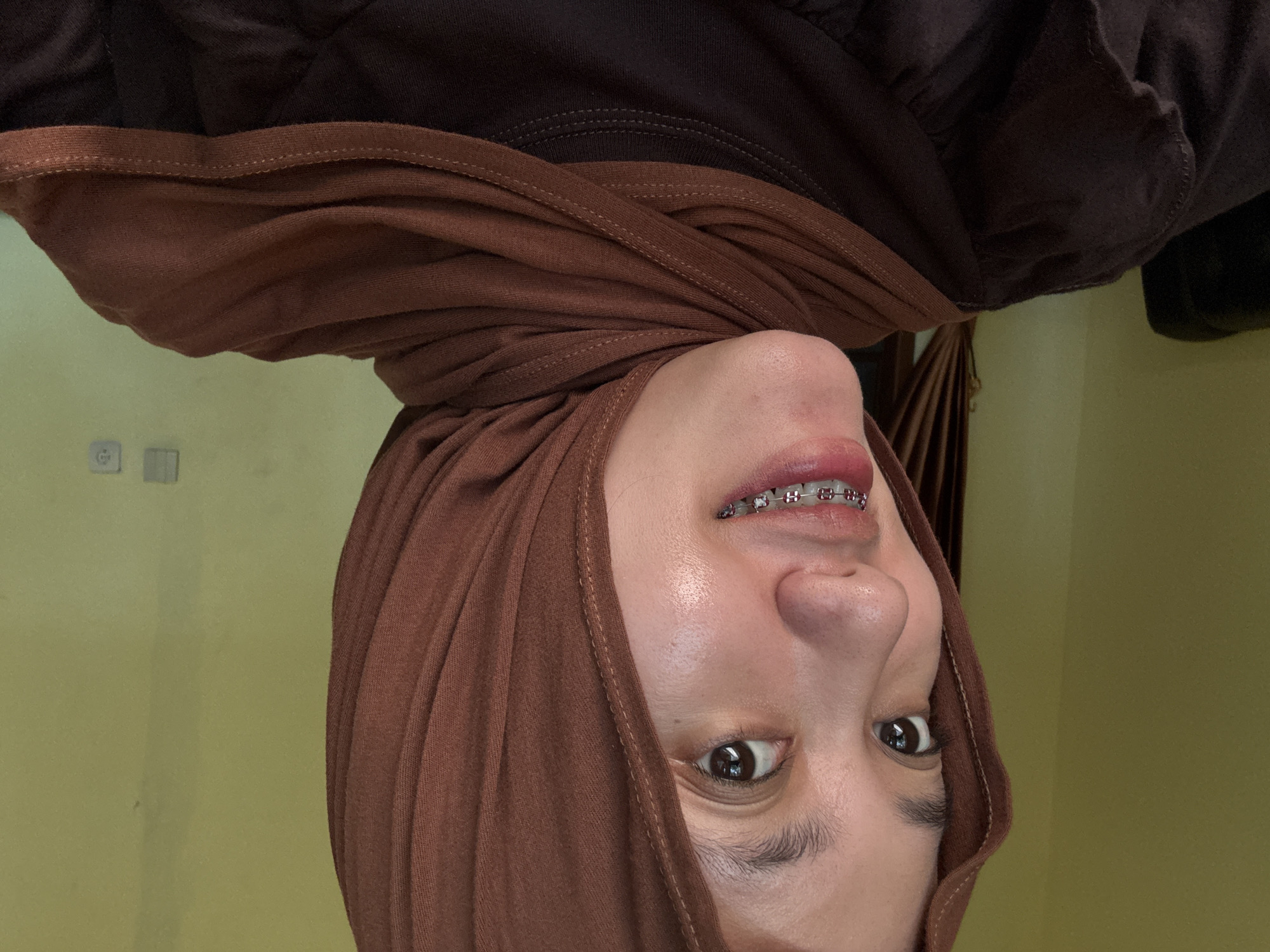 A portrait of a woman is presented upside down. She is wearing a brown hijab. She has braces on her teeth. The background consists of a yellow-green wall and dark brown fabric. The location cannot be confirmed as Gadingrejo, Indonesia.