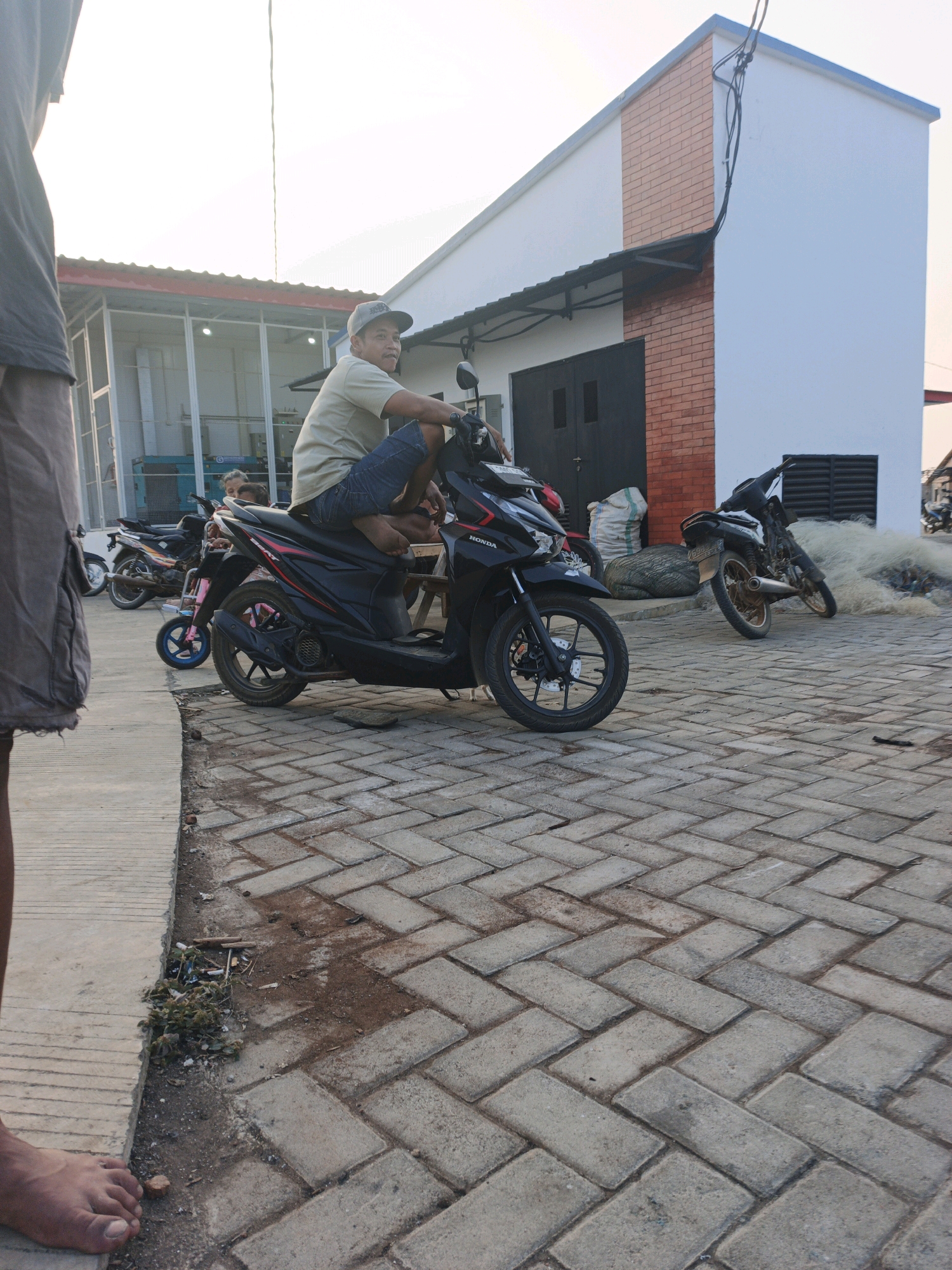 In Bumiharjo, Indonesia, a man wearing a beige t-shirt and blue shorts sits on a black scooter. He is wearing a grey baseball cap and has a relaxed posture. The scooter is parked on a cobblestone pathway in front of a white and red brick building. To the left of the building, there is a glass-enclosed structure. Several other motorcycles are parked in the vicinity, including a smaller pink and blue scooter. In the background, a large pile of white netting is visible near a weathered motorcycle. The scene appears to be outdoors, with a bright sky overhead.