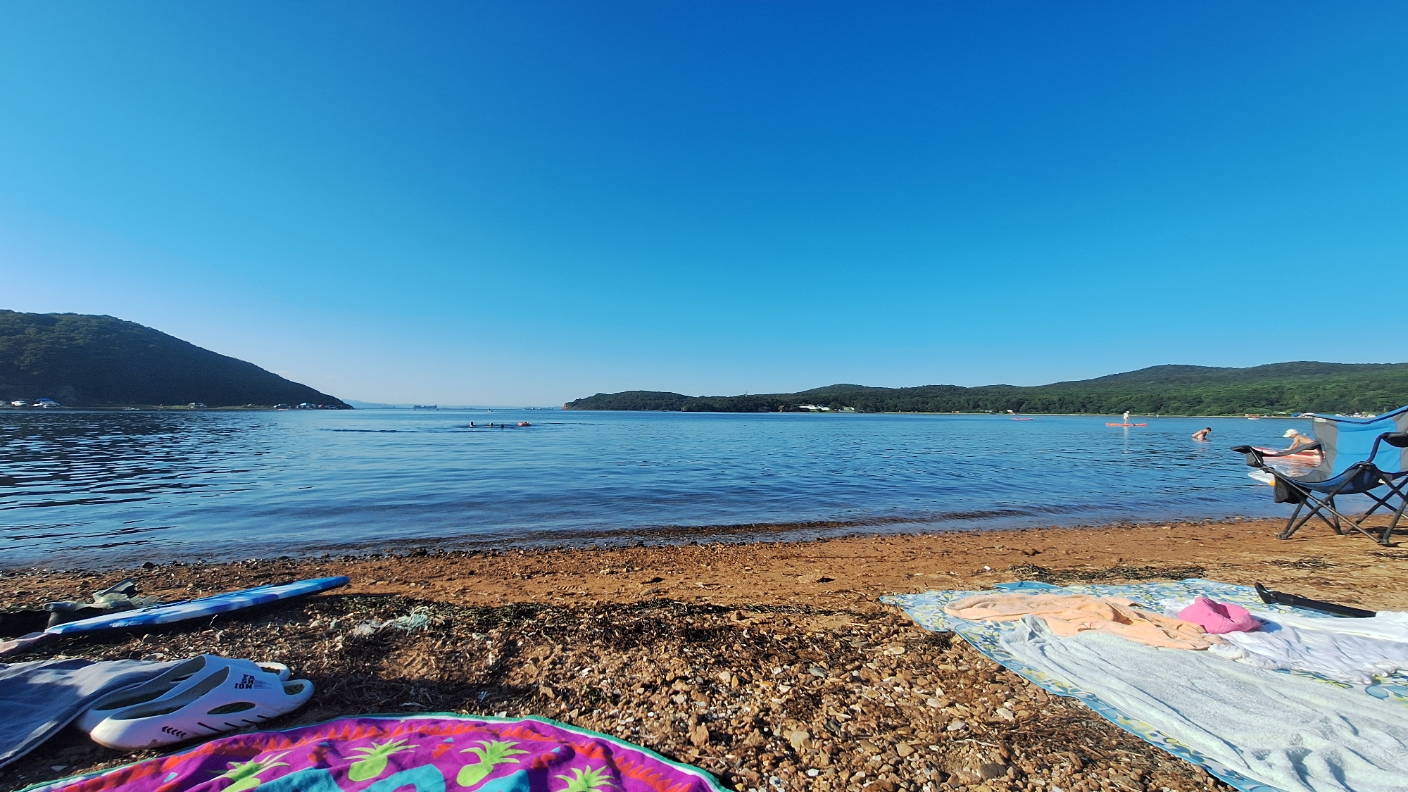 The image depicts a sunny day at a beach, likely on Popova Island, Russia, given the context. The foreground is dominated by a sandy and pebbly shore, with several beach towels and a pair of white sandals spread out. A blue and white fins are also visible. The calm, blue water of the bay stretches out to the horizon, dotted with several people engaged in various water activities. Some are swimming, while others are on paddleboards or in small inflatable boats. In the background, lush green hills rise from the coastline, meeting a clear, bright blue sky. The overall scene conveys a relaxed and recreational atmosphere.