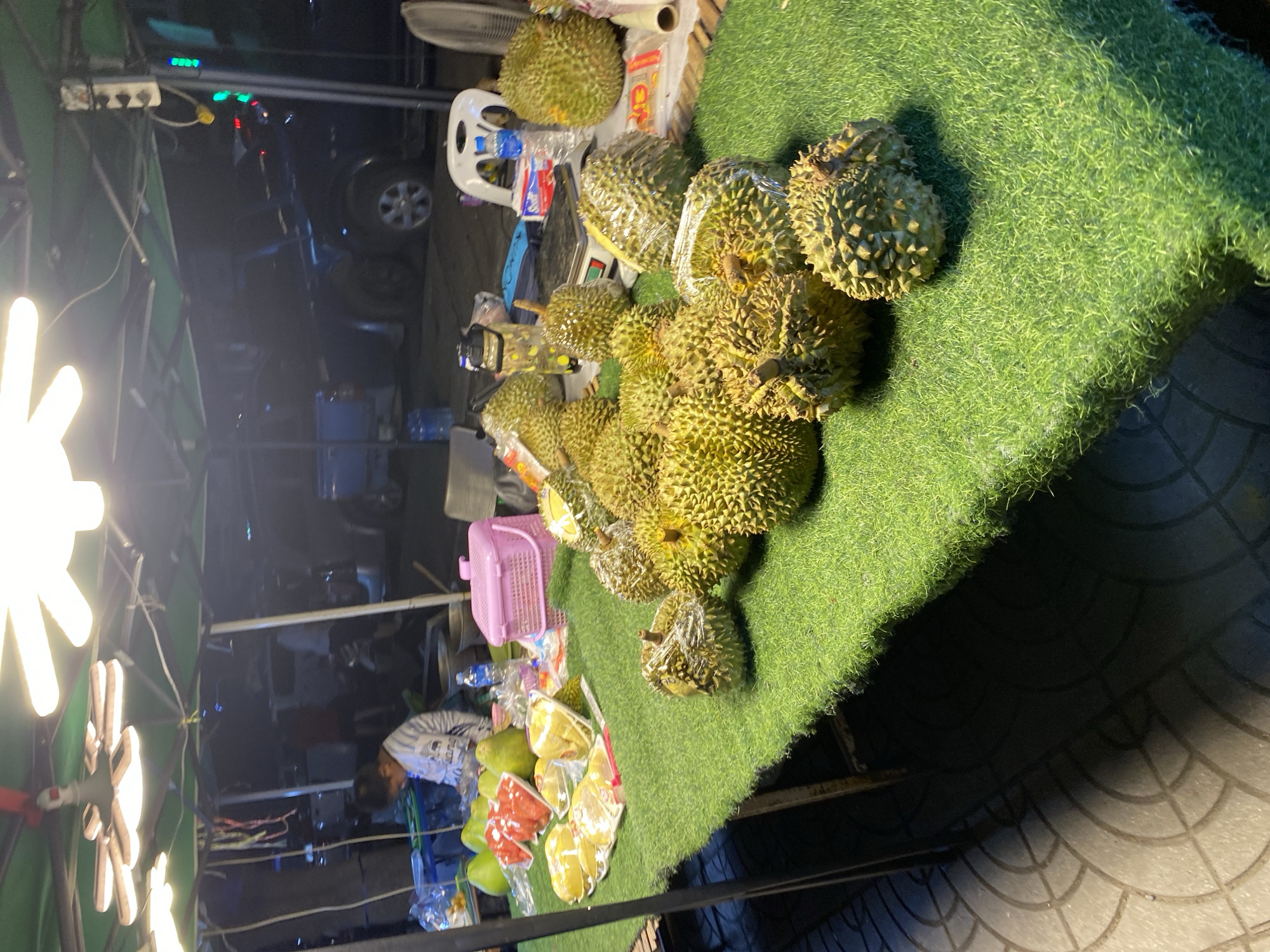 The image displays an outdoor fruit stall at night in Choeng Thale, Thailand, illuminated by bright artificial lights. The central focus is a large collection of durians, many still in their spiky green husks, some wrapped in clear plastic, piled on a surface covered with artificial green grass.
Beyond the durians, various pre-cut tropical fruits are visible in clear plastic bags, including what appears to be watermelon slices and star fruit, alongside some whole green fruits, possibly pomelos. These are also displayed on the grass-covered surface. To the left, behind the stall, a person, likely a vendor, is partially visible sitting down, wearing a striped shirt.
The stall setup includes a pink plastic basket, a white plastic chair (with "PLASTIC" partially visible inverted), a digital scale (partially seen), and a bottle of water with "AQUA PURE" visible on its label. In the background, a large fan is positioned, and further back, a dark street scene with a parked vehicle (possibly a car or truck) is discernible. An electrical power strip with green, red, and blue indicator lights is attached to a structure in the upper left.
The bright overhead lighting casts strong shadows, clearly indicating it is nighttime. The atmosphere suggests a typical bustling night market environment. The ground visible beneath the stall appears to be paved with a patterned surface. No specific weather conditions are discernible, but the outdoor setting is clear. Emotions are not overtly expressed by any visible person.