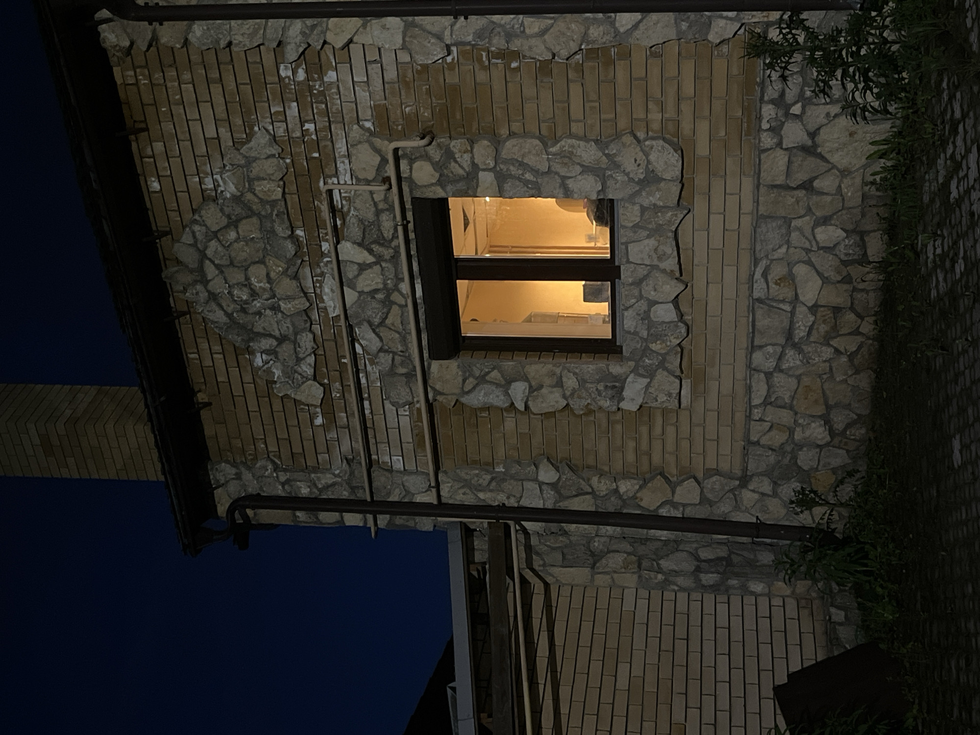 This media file captures the exterior of a building at night in Ореховка, Russia. The building wall features a distinctive alternating pattern of light brown brickwork and irregular grey-brown stone masonry. A prominent rectangular window with a dark frame is centrally located on the wall, emitting a warm, yellow-orange light from the interior, indicating that internal lights are on. Through the window, glimpses of an indoor space with white surfaces, possibly part of a kitchen or utility area, are visible, along with some reflections.
Dark-colored pipes, likely part of a drainage or utility system, run across the stone and brickwork. To the far left of the image, against a deep blue-black night sky, a tall, brick-colored chimney-like structure can be seen. On the right side of the building, dark green foliage or bushes grow along the wall and extend towards a darker, paved ground area. No people are visible, and no specific activity is occurring beyond the illumination from the window. The image clearly depicts a nighttime scene. There is no visible text.