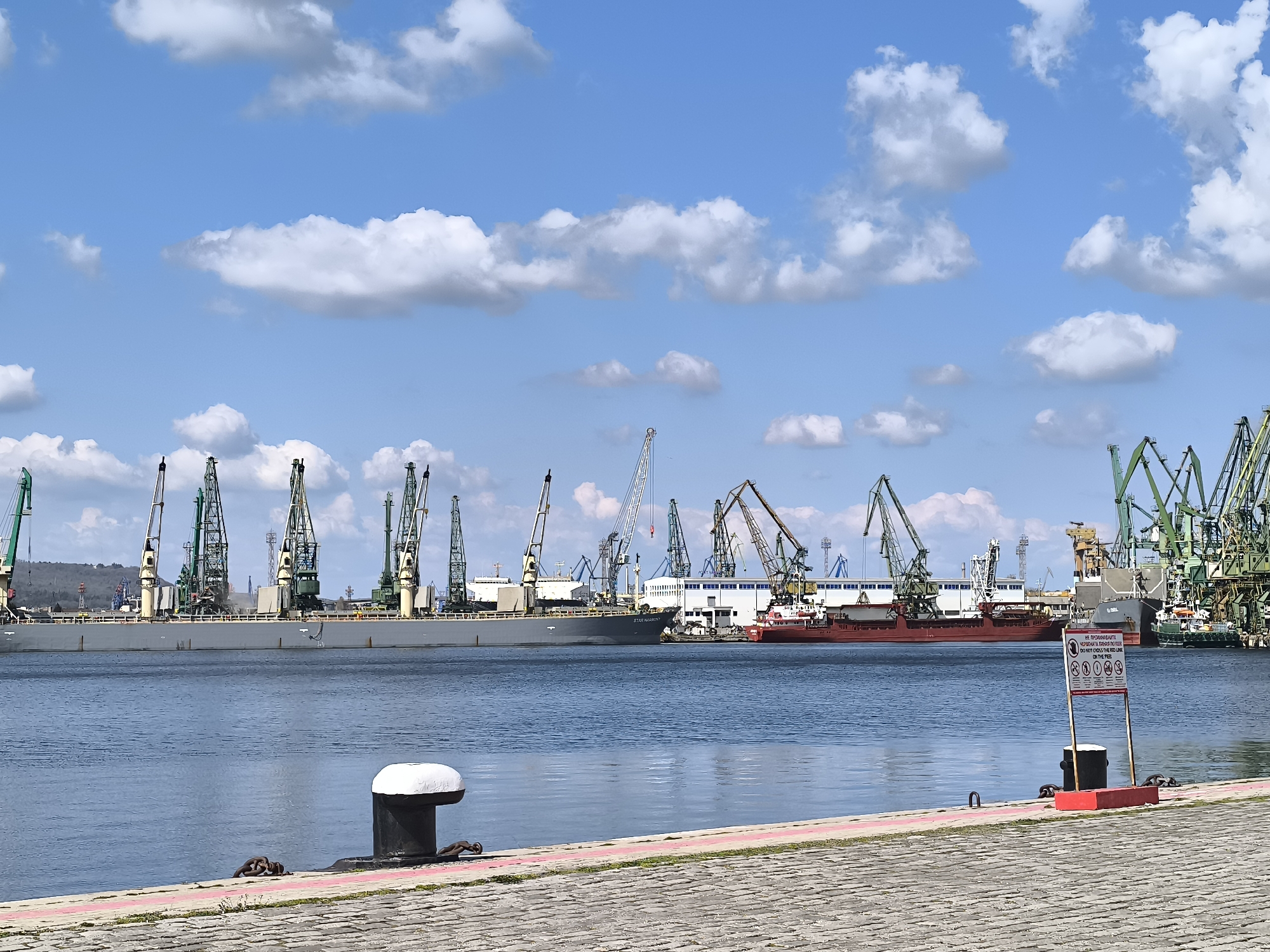The image captures a harbor scene under a blue sky dotted with puffy white clouds. In the foreground, a stone-paved walkway leads to the water's edge, marked by a red line and metal bollards. A sign with text in an unidentified language is placed in front of the water. The middle ground shows the water leading to a port bustling with ships and cranes. The ships are of various colors including grey and red. Numerous cranes, primarily green, gray, and brown, stand behind the ships. The background features structures associated with port operations. The general location is presumed to be an unidentified city.