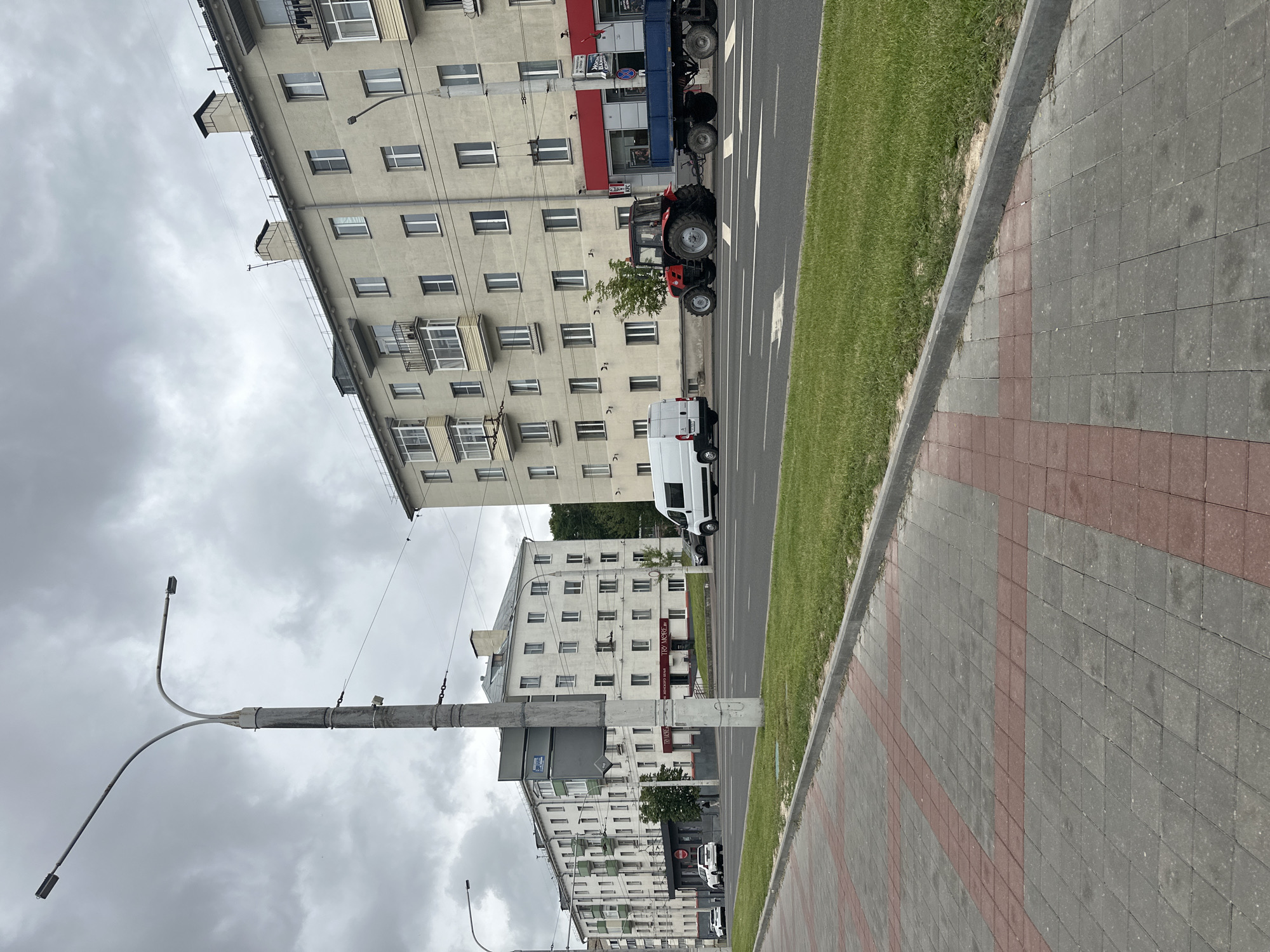 This is a vertical, eye-level, outdoor shot of a street scene in Minsk, Belarus. The image is dominated by tall, light-colored apartment buildings on the left and a paved sidewalk and grassy area on the right. A grey, overcast sky fills the top portion of the frame.
Several vehicles are visible on the street. A red tractor is parked next to a blue trailer truck. Further down the street, a white van is parked. The apartment buildings feature numerous windows and balconies. The sidewalk is paved with grey and red interlocking bricks.
No people are clearly visible in the image, and the overall mood appears neutral, likely due to the overcast weather. The scene depicts an urban environment during daylight hours. No prominent text is legible.