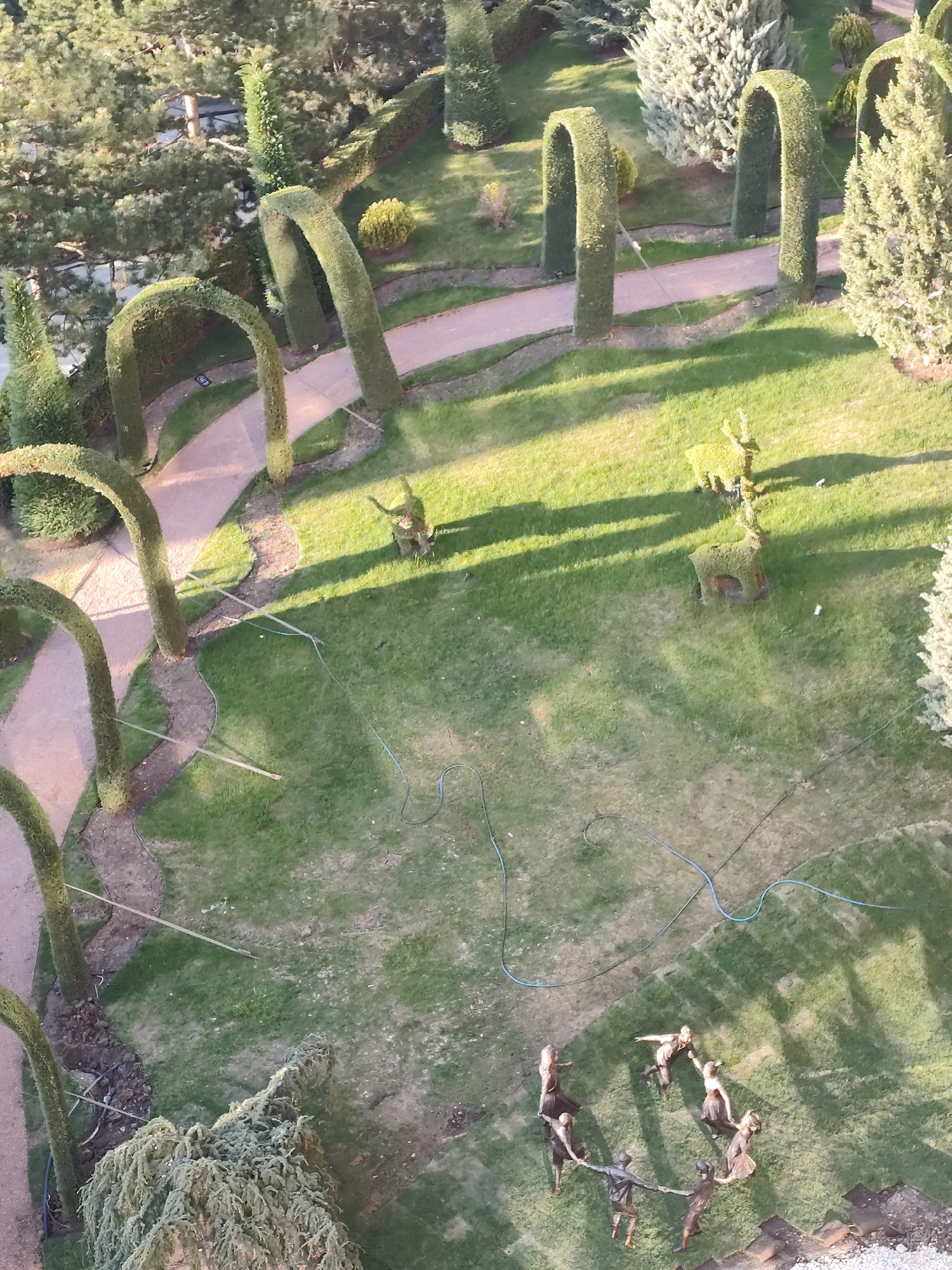 An aerial view captures a meticulously maintained formal garden or park, bathed in the warm light of either early morning or late afternoon, as evidenced by the long shadows stretching across the lawn. The landscape features a large expanse of vibrant green grass, crisscrossed by winding paved pathways.
A prominent element of the design is the series of tall, gracefully curved topiary arches, crafted from neatly trimmed hedges, that frame sections of the pathways. Various trees and shrubs, including dense conifers and lighter-colored evergreens, are interspersed throughout the grounds, adding depth and texture.
On the central grassy area, at least two topiary sculptures shaped like deer or similar horned animals are visible, casting distinct shadows. Towards the bottom of the image, a group of approximately seven bronze or dark-colored statues, appearing to be children, are depicted holding hands in a circle on the grass, suggesting a dance or playful activity. Their dynamic poses convey a sense of joy and movement.
Several blue and black hoses or cables are laid across the grass, possibly for irrigation or maintenance. There is no visible text in the image. The scene depicts a tranquil and artfully designed recreational space, located in Simeiz, Ukraine.