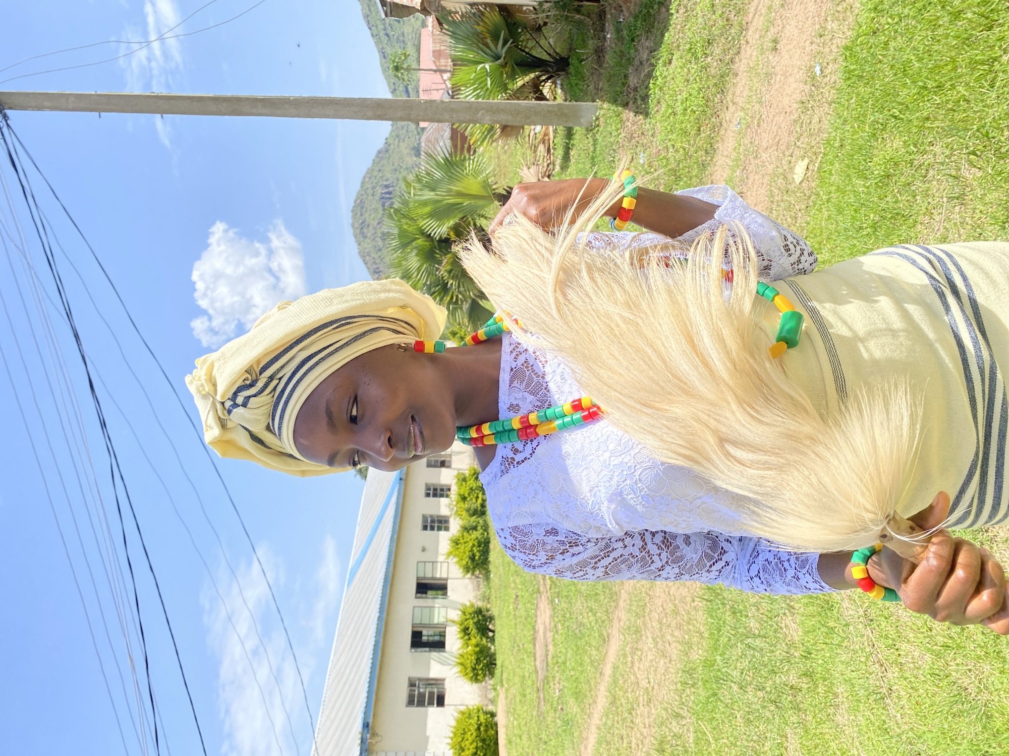 A person is shown from the chest up, facing left and looking downwards. They are wearing a yellow headscarf with dark stripes and a white lace top. Around their neck is a colorful beaded necklace with green, yellow, and red beads, matching a bracelet on their right wrist. In their right hand, they hold a large fan made of light-colored, wispy material. The person has a bright, cheerful expression.
The background suggests an outdoor setting under a clear blue sky with fluffy white clouds. Green grass covers the ground, and in the mid-ground, there is a building with a light-colored exterior and blue trim, featuring multiple windows. Palm trees and other greenery are visible near the building, and a lush, green mountainside rises in the far background. The presence of utility poles and wires, along with the style of the building, might indicate a semi-urban or institutional location. The lighting suggests it is daytime, likely sunny. The overall mood is pleasant and vibrant. The location context provided is Vom, Nigeria.