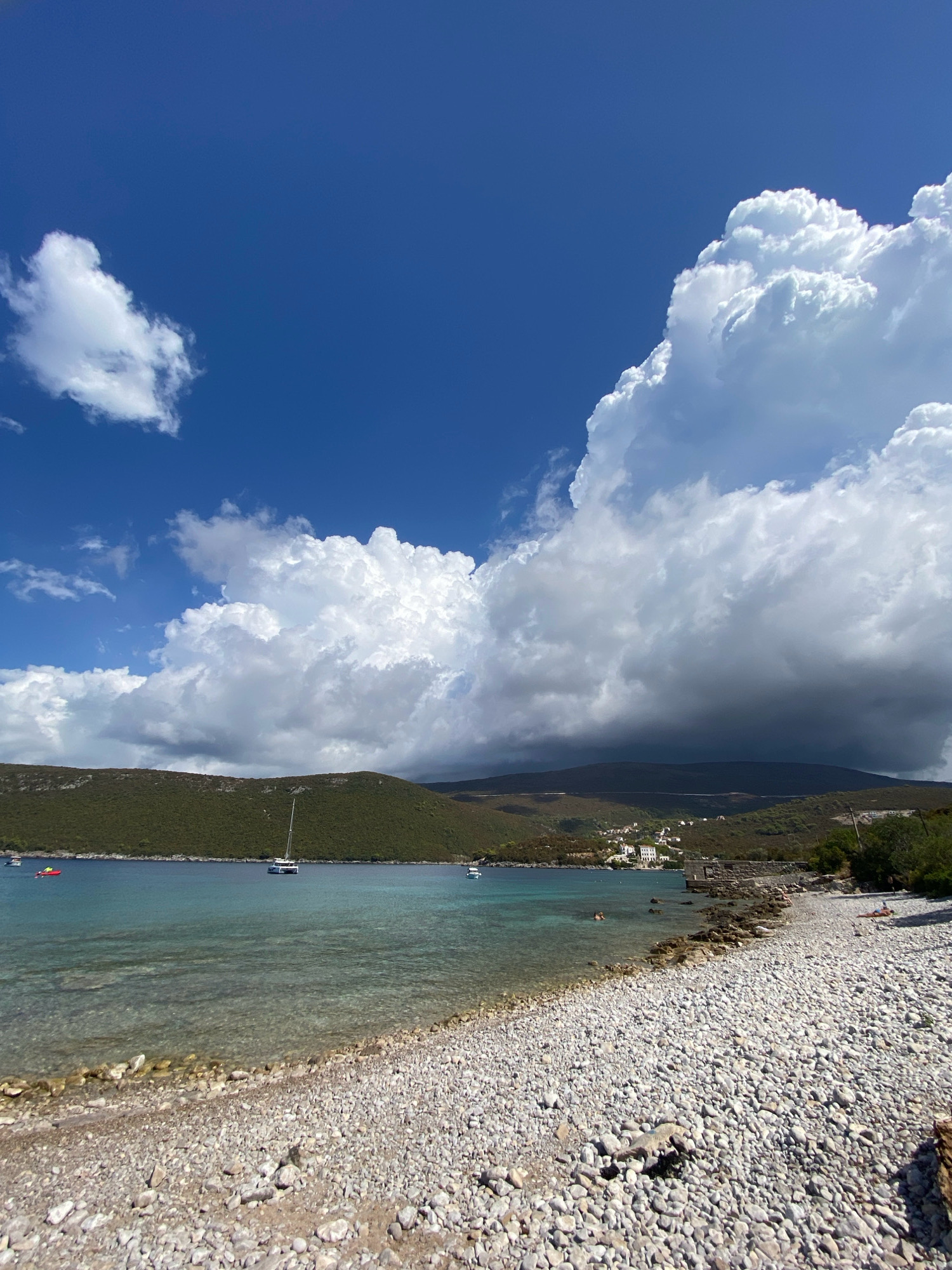 The image depicts a serene coastal scene in Luštica, Montenegro. The foreground is dominated by a pebble beach, with numerous smooth, grey stones stretching towards the clear, turquoise water. The water itself is calm and shallow near the shore, gradually deepening as it recedes towards a verdant, tree-covered hillside that forms the middle ground. Several boats are visible on the water, including a white catamaran with its sail raised, anchored further out. A small red boat is closer to the shore. The background features rolling hills that recede into the distance, partially obscured by dramatic, billowy clouds. These clouds are a prominent feature of the sky, with a large cluster of white cumulonimbus clouds dominating the upper right portion of the frame, casting a shadow over the distant hills. The sky is a vibrant blue, with smaller, wispy clouds scattered across it. A few individuals are visible on the beach and in the water. One person is lying on a towel on the beach, sunbathing. Another person is swimming in the water. A cluster of white buildings, characteristic of a small village, is nestled on the slope of the hill. The overall impression is one of a picturesque and tranquil summer day in a Mediterranean coastal location.