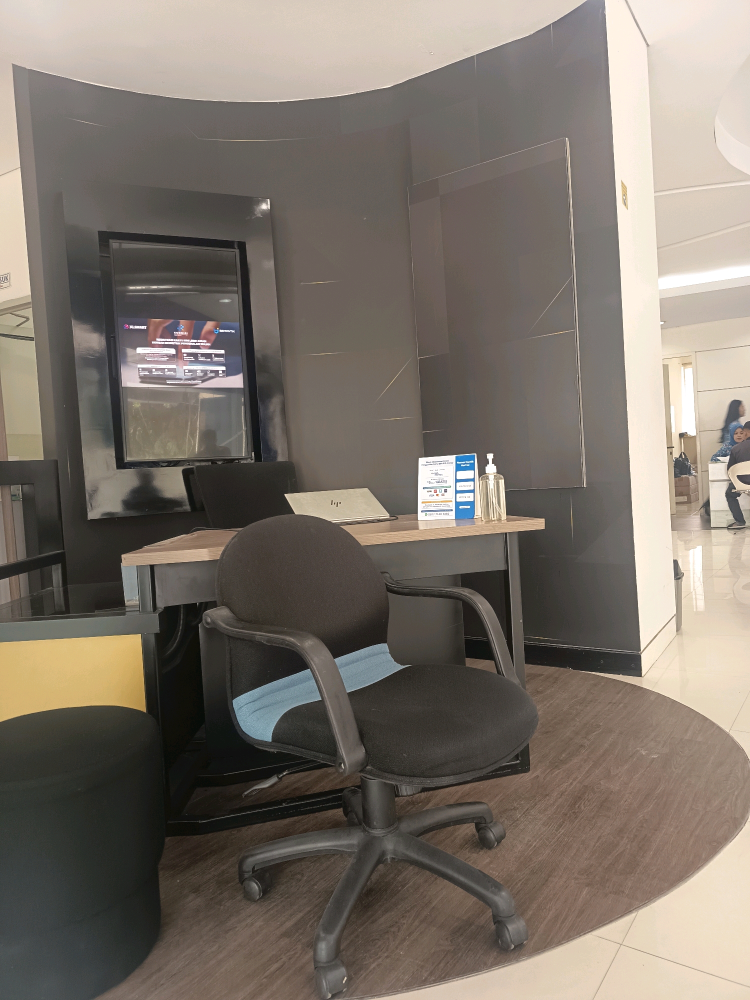 The image depicts an indoor scene, likely a business or service area, with a focus on a desk and office chair. The desk is made of wood with a metal frame and has a laptop placed on it, along with a blue brochure and a clear hand sanitizer dispenser. The chair is a black office chair with wheels and a light blue accent on the backrest. To the left of the desk, a black cylindrical ottoman is visible. Behind the desk, a large, dark gray tiled wall serves as a backdrop, with a mounted flat-screen display showing graphics. The flooring consists of light-colored tiles and a circular wooden mat under the desk and chair. In the background, through an opening, a brighter area is visible with people, suggesting a waiting area or customer service counter. The overall lighting is moderate, with a slight glare on the tiled walls. There is no direct evidence in the image to suggest the location is the city of Bogor, Indonesia.