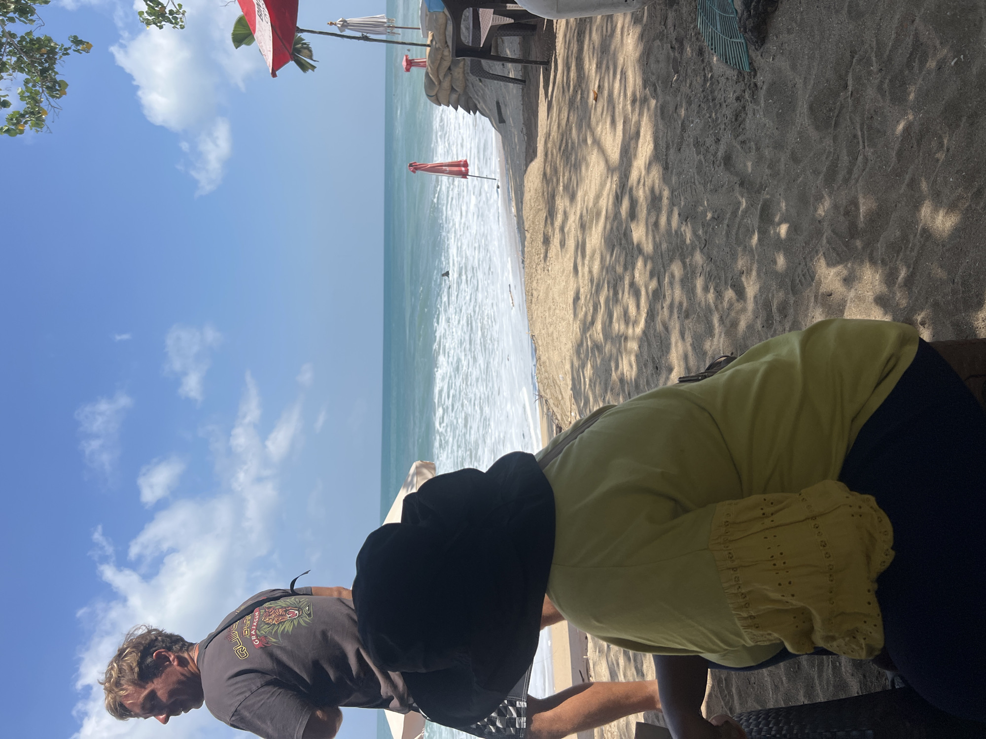 The image captures a beachfront scene in Kuta, Indonesia, with the ocean and sky dominating the background. In the foreground, a person wearing a black bucket hat and a yellow shirt is seated, with their back to the camera. To their left, a man in a dark t-shirt stands, facing away from the camera and looking towards the ocean. The beach sand is visible, with shadows from overhead foliage cast upon it. In the midground, the ocean exhibits gentle waves and a sandy shore. Several beach umbrellas, some red and some white, are present along the coastline, along with sandbag barriers. Two chairs and a table are situated on the sand to the right of the frame. The sky is blue with scattered white clouds.