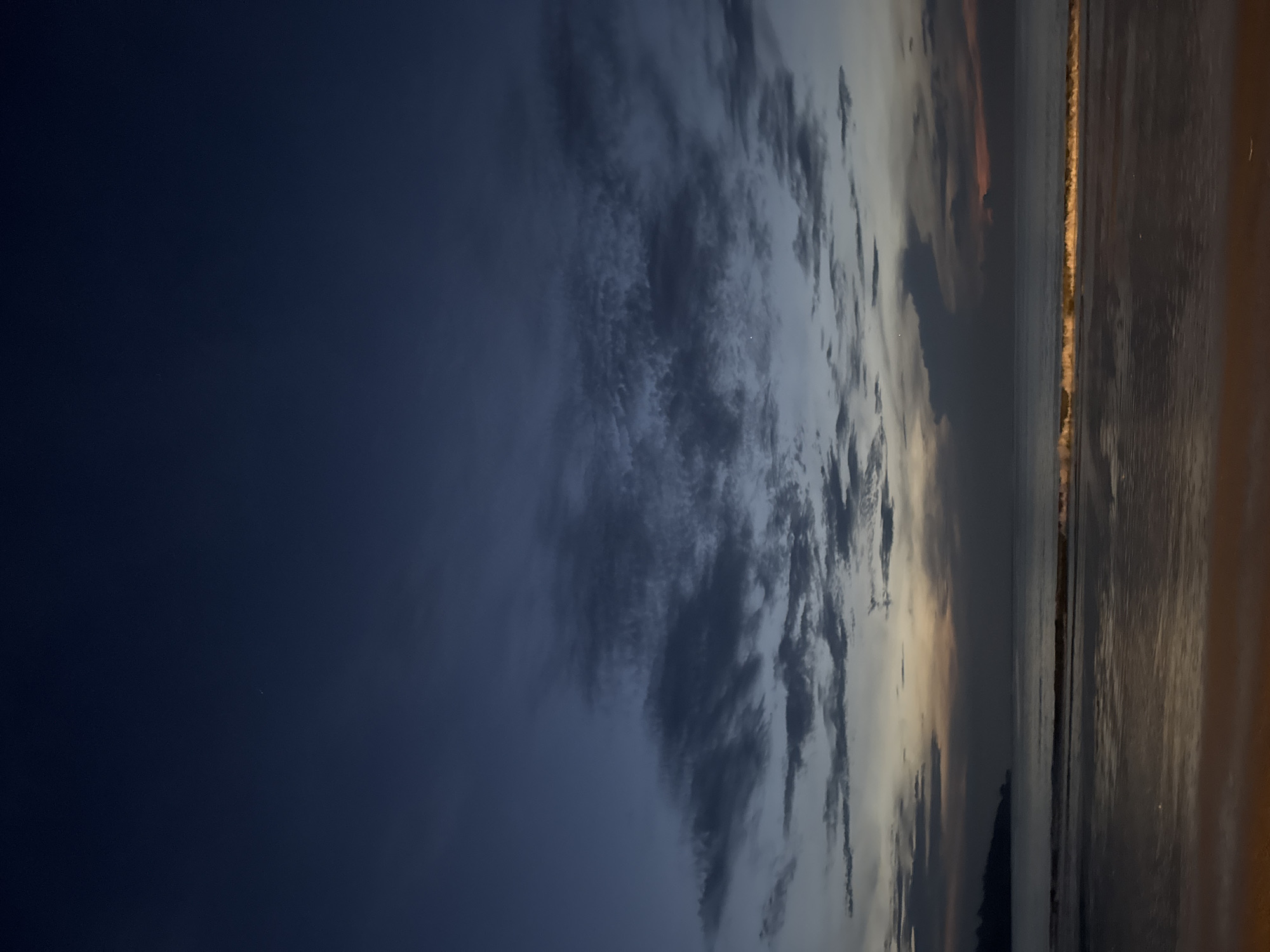 The image depicts a coastline at dusk, likely in Kuta Selatan, Indonesia. The left side of the frame features a deep blue sky transitioning into a cloudscape with varying shades of gray and hints of orange towards the horizon. The right side shows a body of water, reflecting the light from the sky. A thin strip of land or beach is visible between the water and the sky, with a line of lights along it, possibly indicating a settlement or infrastructure. The image is oriented vertically.