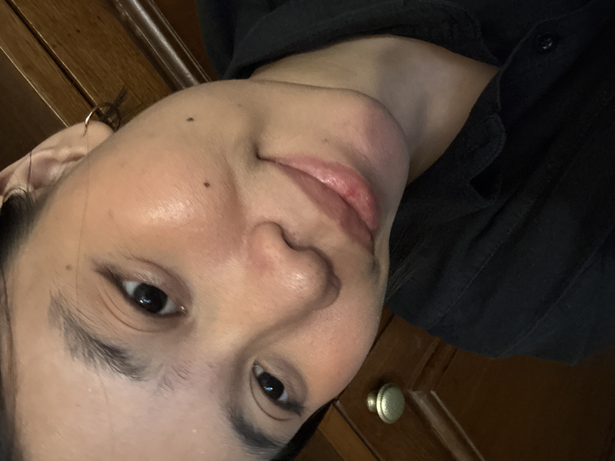 A close-up, self-taken photograph features a woman with medium-toned skin, dark eyes, and dark eyebrows, looking towards the camera with a slight smile. She is wearing a black collared shirt. Her right ear has a small hoop earring. There is a mole on her cheek and another below her lip. The background consists of wooden furniture, likely a dresser, with a brass drawer pull visible on the left side. The lighting suggests an indoor setting.