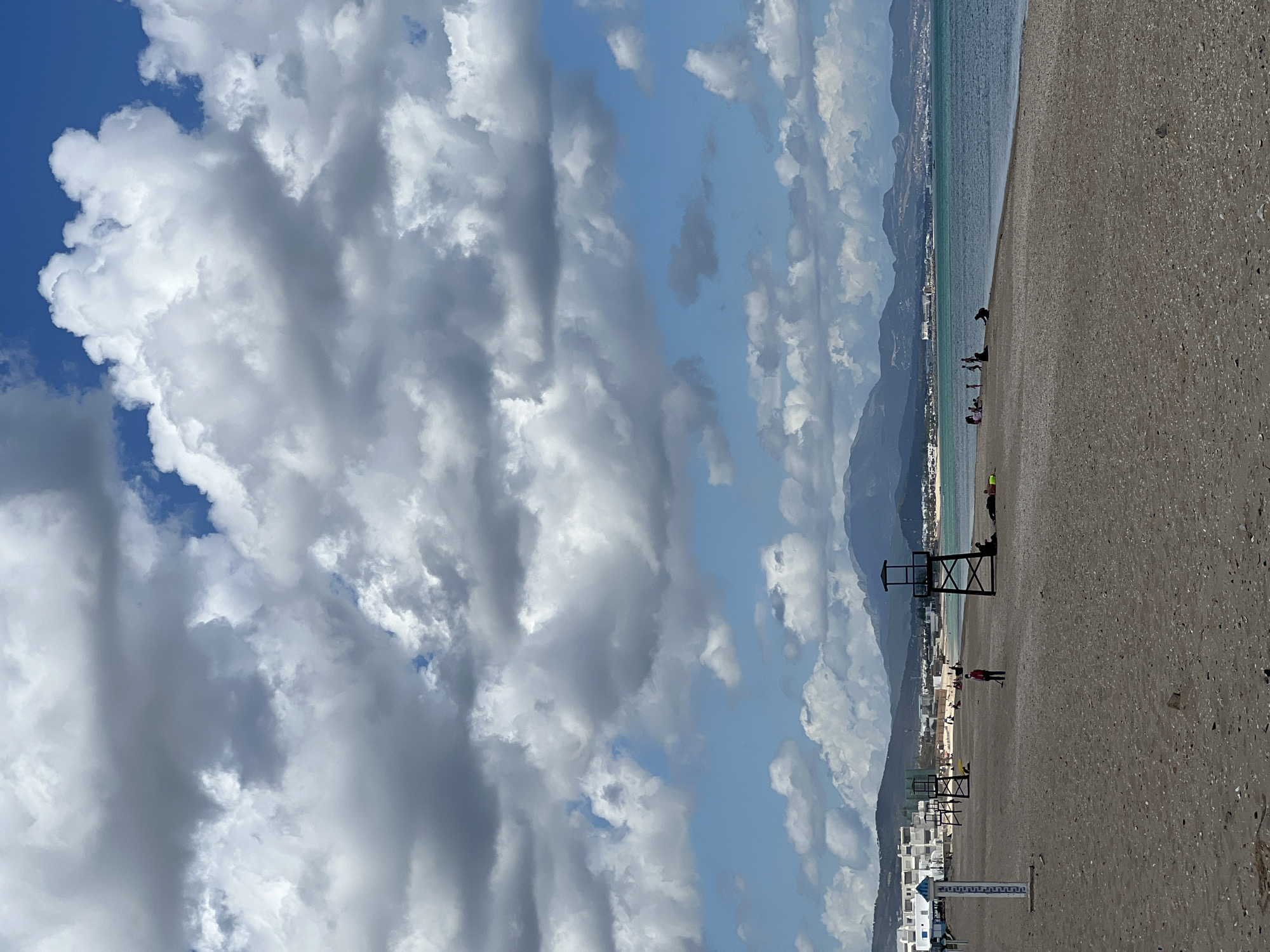 The image depicts a beach in M'Diq, Morocco, under a sky filled with cumulus clouds. The beach appears to be dark sand, and the sea is visible along the top right edge. Mountains are visible in the background, across the bay.
There are several lifeguard towers, some occupied. There are also people on the beach, both near the shoreline and further back. The town is seen in the background with white buildings.