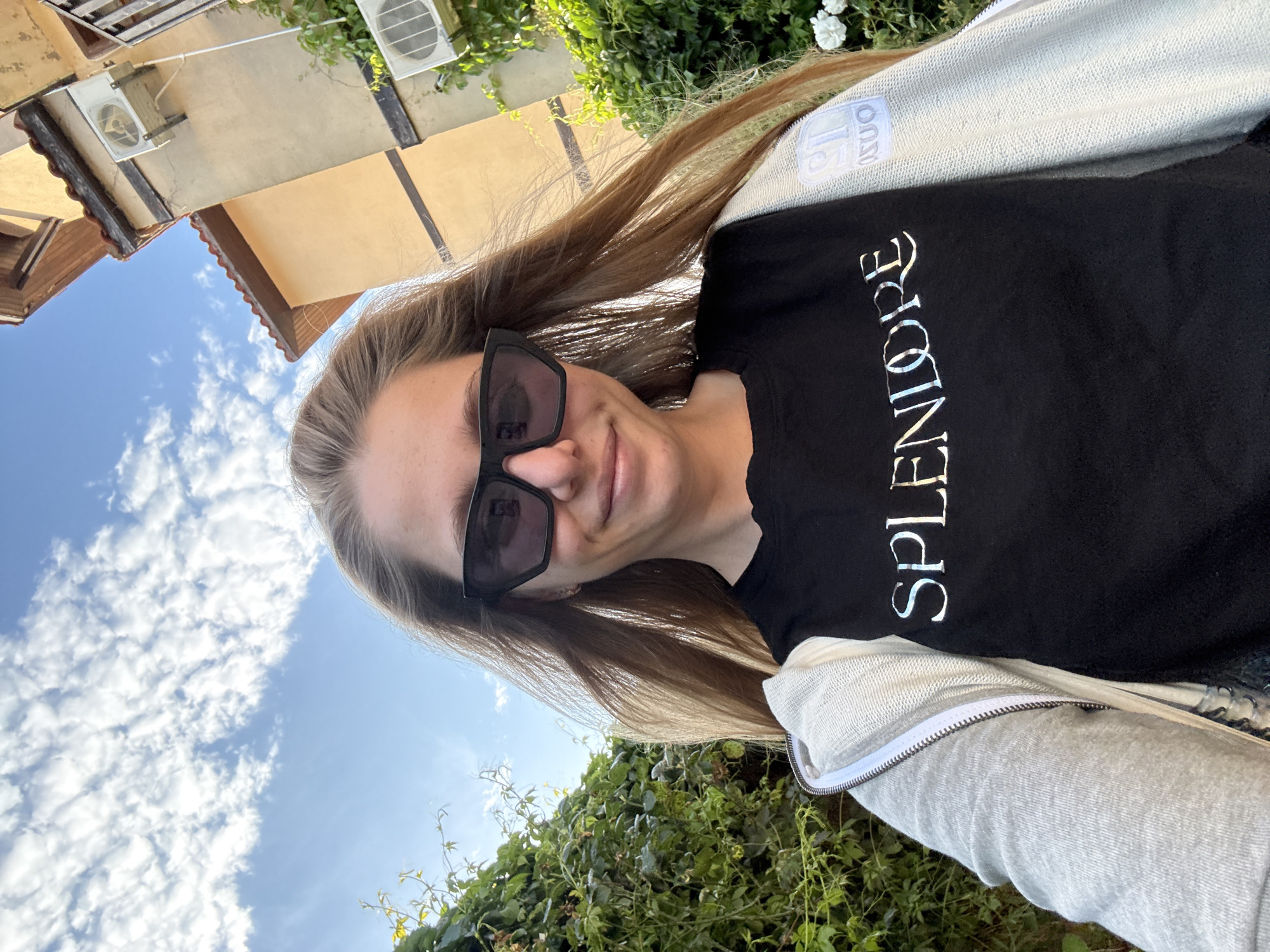 A young woman is taking a selfie outdoors. She is wearing sunglasses and a black t-shirt with the word "SPLENDORE" in silver lettering. She has long, blonde hair that is being blown by the wind. Behind her, there is a building with air conditioning units and a clear blue sky with scattered clouds. Lush green foliage is visible in the foreground. The image appears to have been taken on a sunny day. The woman has a slight smile on her face, suggesting a relaxed and happy mood. The building and the foliage suggest an outdoor setting, possibly a warm climate.