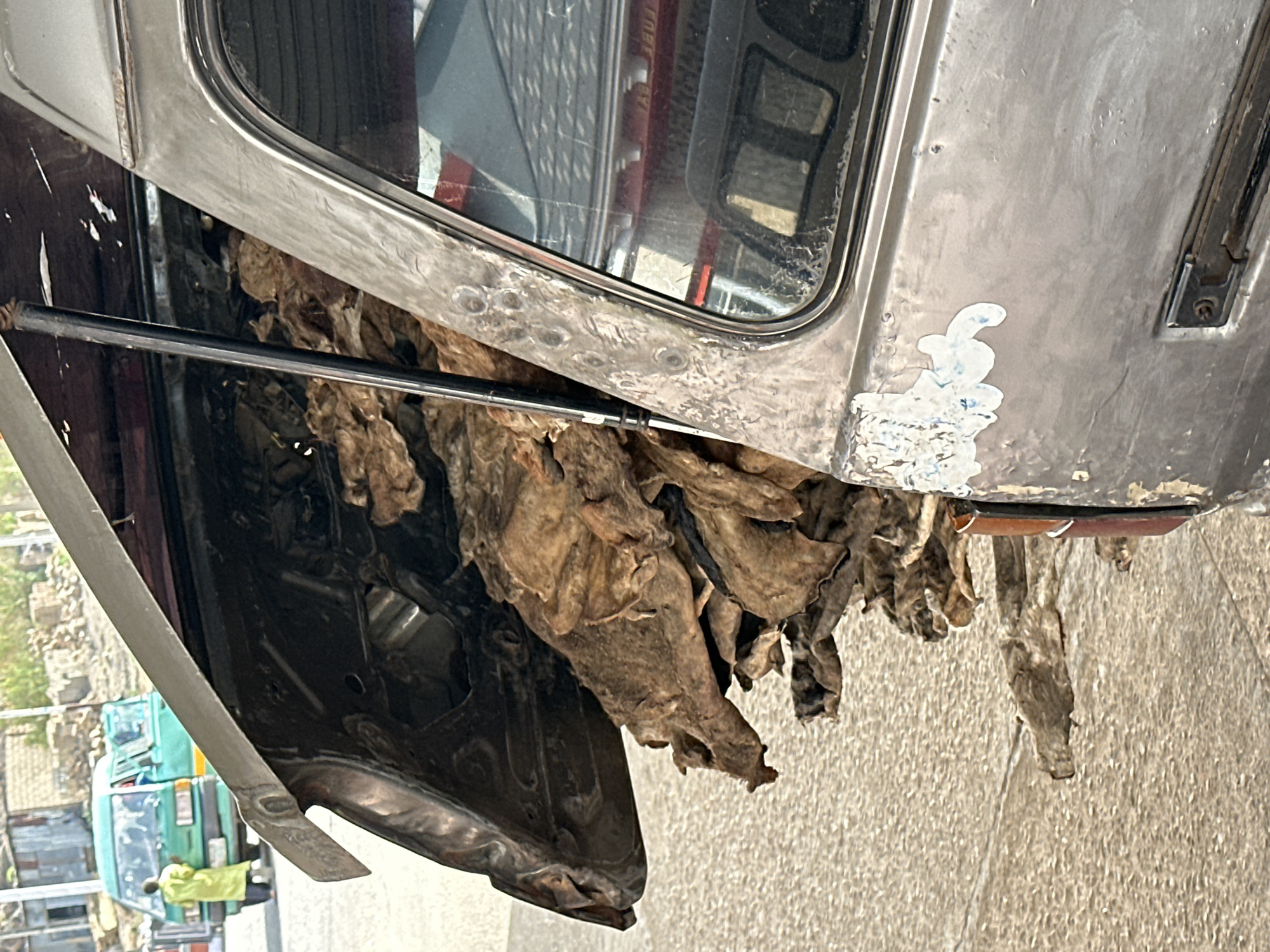 The image depicts the underside of a damaged vehicle, possibly a bus or truck, with exposed insulation hanging down. The vehicle is silver/grey, and the insulation is brown and tattered. A black metal bar extends across the vehicle's underside. The background is blurred, but a green vehicle and buildings are visible, suggesting an urban environment. Based on the prompt, this scene is located in Jos, Nigeria.
