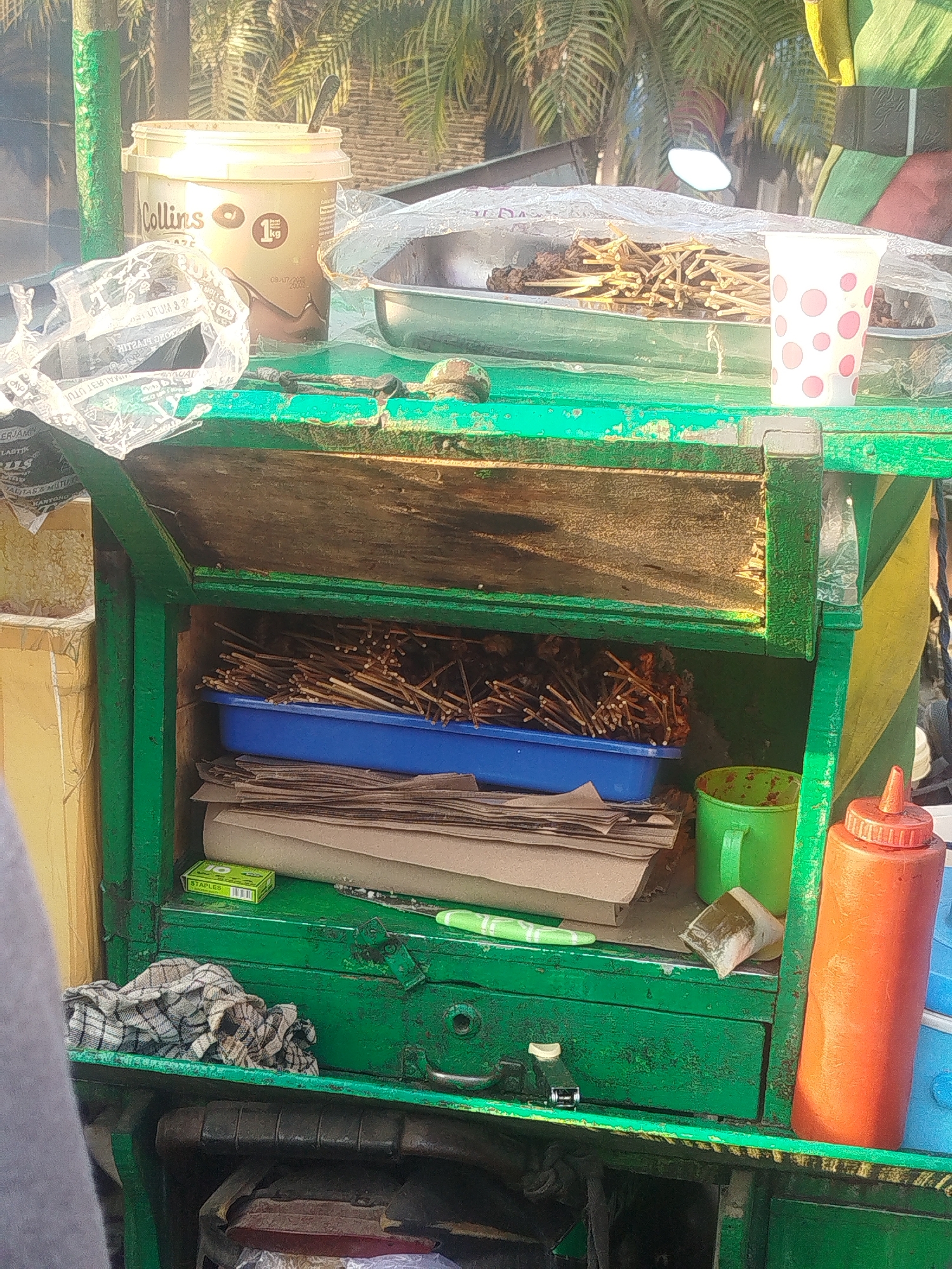 This image shows a close-up of a food cart in Jambu, Indonesia. The cart is green and appears to be made of wood and metal. In the foreground, a person's arm and shoulder are visible on the left, wearing a gray long-sleeved shirt. The cart has several compartments. The top compartment contains a large white tub with the brand "Collins" and "1 kg" printed on it, along with a spoon. Next to it, a tray holds skewers of food, possibly meat or satay, covered in plastic wrap. A pink and white polka-dotted paper cup sits beside the tray.
Below this, a larger wooden compartment opens to reveal a blue plastic tray filled with more skewers. Beneath the blue tray, stacks of brown paper and a green box labeled "STAPLES" are visible. To the right of these, a green plastic cup and a small square object with a brown substance on it are present. Further to the right, an orange squeeze bottle, likely for ketchup or sauce, stands upright. The background shows palm trees and a building, suggesting an outdoor setting. The cart is weathered, with paint chipping in several places, indicating use. The lighting suggests it is daytime, possibly late afternoon due to the warm tones.